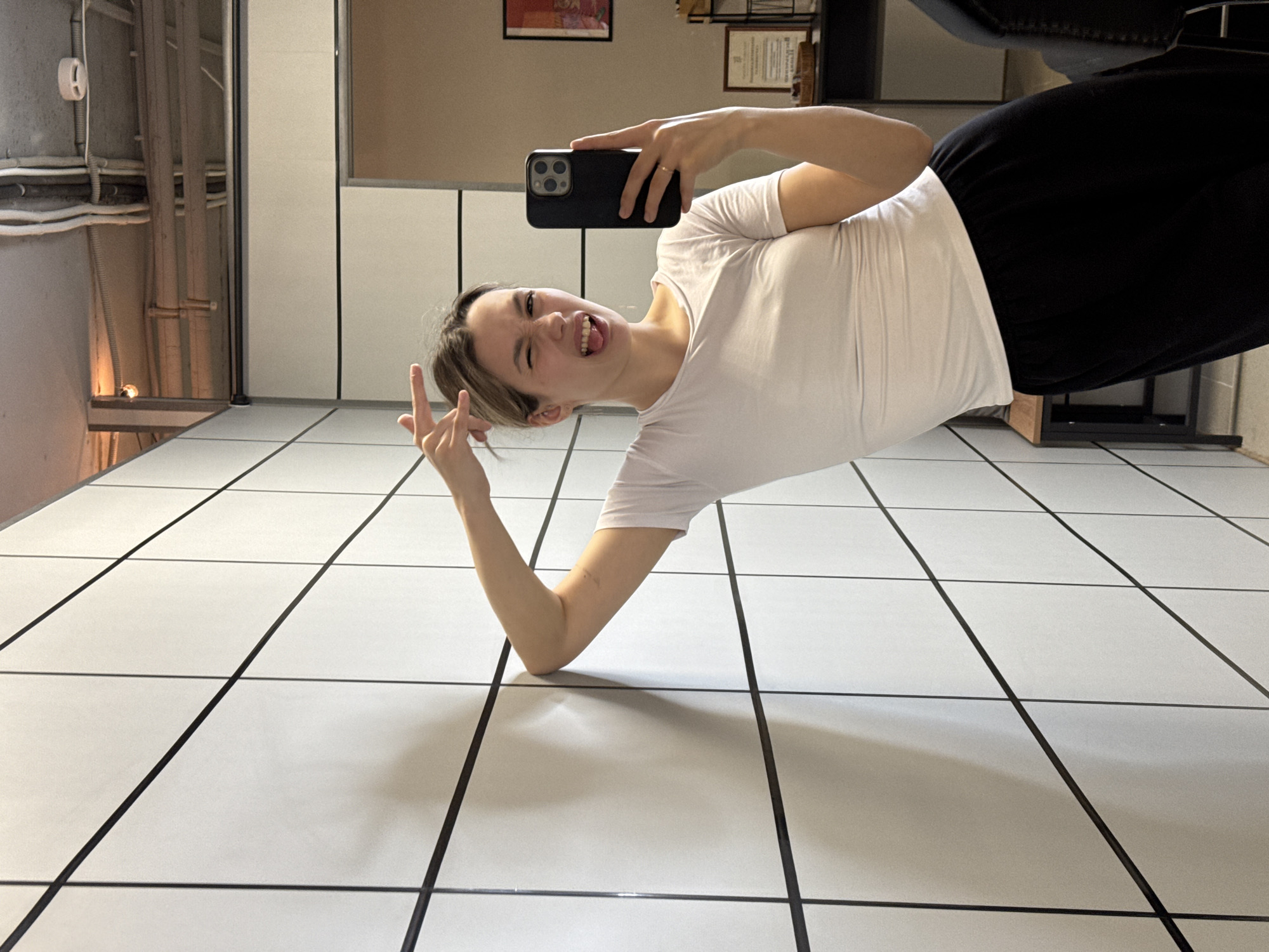 A young woman is depicted in a playful selfie, rotated 90 degrees clockwise, giving the impression that she is lying on a vertical surface. She has light brown hair and is wearing a white t-shirt and black pants. With a mischievous expression, she squints her eyes, sticks her tongue out slightly, and forms a 'V' sign with her left hand. Her right hand holds a black smartphone, likely an iPhone, with a triple camera lens, which she is using to capture the image. A ring is visible on her right hand's ring finger.
The setting is indoors, featuring prominent white square tiles with thick black grout lines, creating a distinct grid pattern that extends across the floor and up what would typically be a wall. In the background on the left, there are exposed pipes and conduits, suggesting an industrial or functional part of a building. To the right, framed artwork or certificates hang on a wall, and a dark piece of furniture, possibly a chair or couch, is partially visible. The overall mood is lighthearted and humorous. There is no visible text.