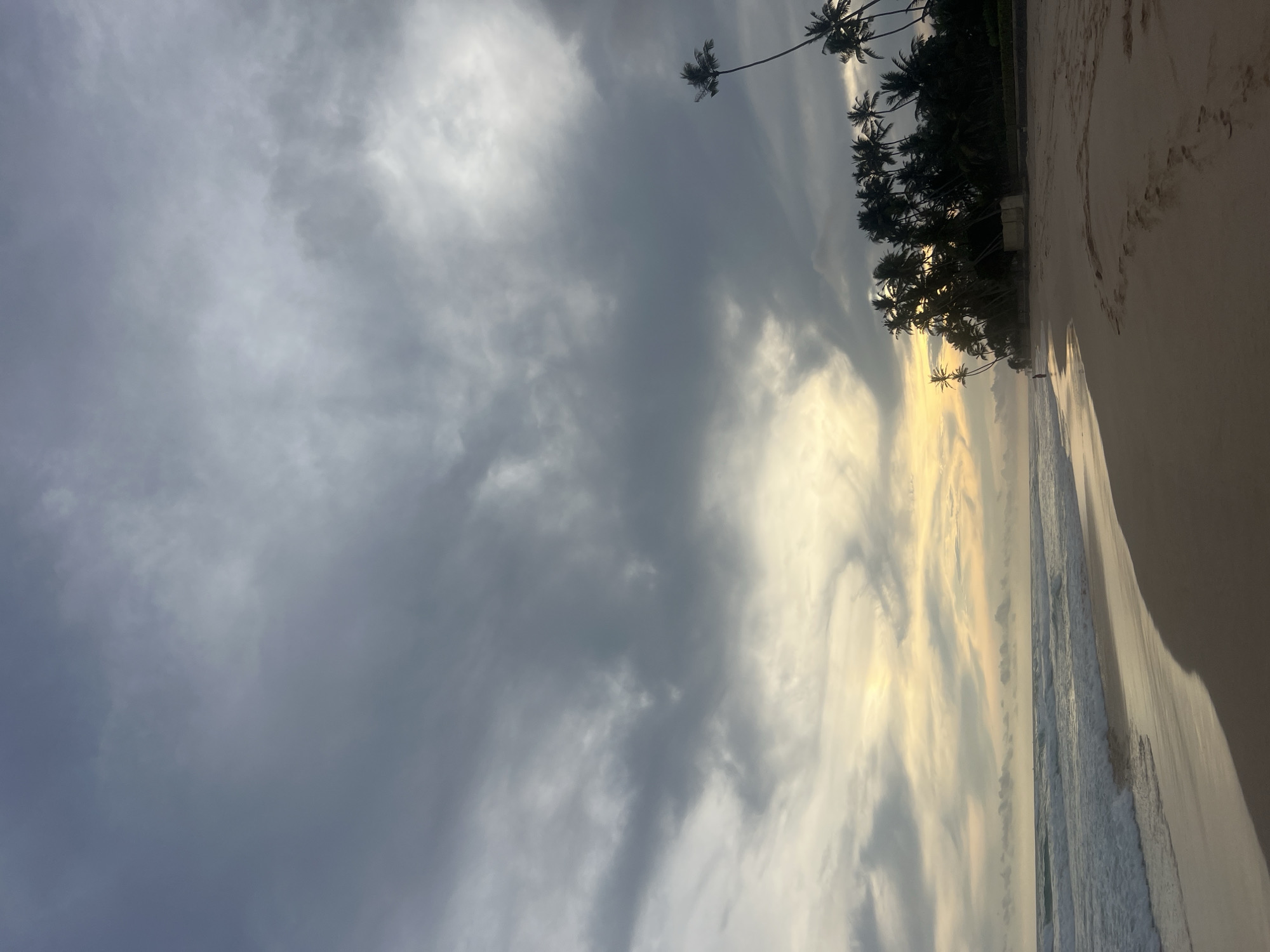 This image, rotated clockwise, captures a serene tropical beach scene, likely at sunrise or sunset in Habaraduwa, Sri Lanka. The upper portion is dominated by a dramatic cloudy sky; darker grey clouds mass on the left, transitioning to lighter, more ethereal formations on the right where a warm, golden light breaks through, illuminating the horizon and reflecting softly on the ocean.
A wide expanse of light-colored sandy beach occupies the lower right quadrant, showing subtle undulations. To its left, gentle ocean waves lap at the shore, creating delicate white foam. A dense line of tall palm trees forms a dark silhouette along the beach's edge, their fronds occasionally extending into the cloudy sky. Partially obscured by the foliage, some light-colored structures, possibly resort buildings, are visible among the trees. A very small, distant figure can be seen walking leisurely along the waterline, adding a sense of scale to the vast landscape. The overall mood is tranquil yet awe-inspiring, with the interplay of light and shadow under the clouded sky. No visible text is present.