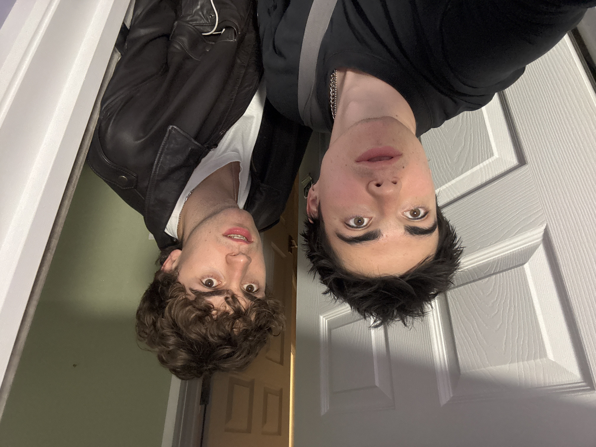 Two young men are positioned upside down in front of two different doors. The man on the left has curly brown hair, wears a white t-shirt under a black leather jacket, and has a silver chain visible around his neck. The background on the left is a light green wall and a wooden door. The man on the right has dark hair and wears a black long-sleeved shirt with a silver chain around his neck. The background on the right is a white paneled door. The image does not contain sufficient information to determine its location in London.