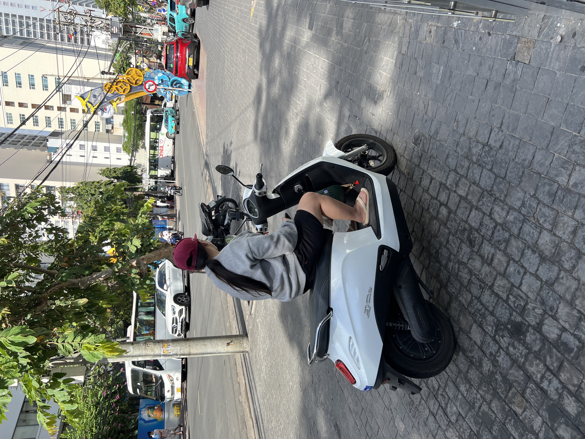 In Nha Trang, Vietnam, a person wearing a maroon baseball cap and a grey sweatshirt is seated on a white electric scooter, facing away from the camera. They are wearing black shorts and light-colored flip-flops. The scooter has a black seat and black tires, with "EVO Lite" visible on its side. The scooter is parked on a cobblestone sidewalk next to a street with vehicular traffic. In the background, a white bus with "ANEX" written on it is visible, along with several cars, including a red VinFast car. Buildings line the street, and overhead electrical wires crisscross the sky. Trees with green leaves provide some shade. The lighting suggests it is daytime.