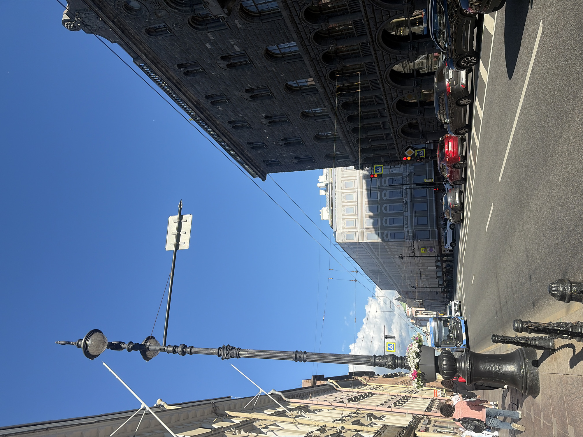 The image depicts a street scene in Saint Petersburg, Russia, on a bright, clear day. The perspective is from a sidewalk looking down a wide, paved street with multiple lanes of traffic. The architecture on both sides of the street is grand and historic, characteristic of Saint Petersburg.
On the left, a building with a light-colored facade, possibly stuccoed, features ornate detailing and tall windows. A black lamppost with decorative elements stands prominently on the sidewalk, adorned with a large planter overflowing with flowers. People are walking on the sidewalk, some facing away from the camera, carrying backpacks.
On the right, a dark stone building dominates the view, displaying arched windows and detailed stonework, suggesting a significant historical structure. Overhead wires crisscross the sky, indicating trolleybus or tram infrastructure, common in Saint Petersburg.
The street itself is active with vehicles. A blue and white trolleybus is visible moving forward, and several cars, including a red sedan, a dark SUV, and a black SUV, are driving in the same direction. Traffic lights are visible, one showing red. Pedestrian crossing signs are also present. The sky is a vibrant blue with a few wispy clouds. The overall impression is of a bustling urban environment within a historically rich city.