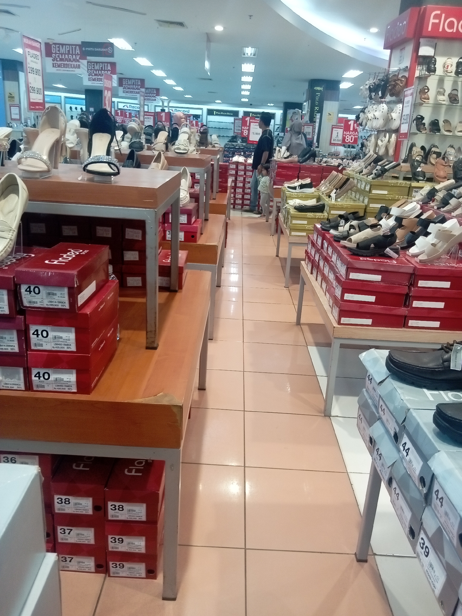 The image depicts an indoor shoe store, likely within a mall in Batam, Indonesia, given the signage. The foreground shows several display tables with stacks of red shoe boxes, some with size labels like "40" and "38". On top of these boxes, various women's shoes are displayed, including high heels and sandals. To the right, a metal shelving unit is filled with more shoes. A person, a man in a dark shirt and jeans, is visible in the mid-ground, standing near a display of shoes, with a child standing beside him. The background reveals more shoe displays and store signage, some with promotional text such as "GEMPIT SEMARAK KEMERDEKAAN". The floor is tiled, and fluorescent lighting illuminates the store. The overall scene is one of a retail environment focused on selling footwear.