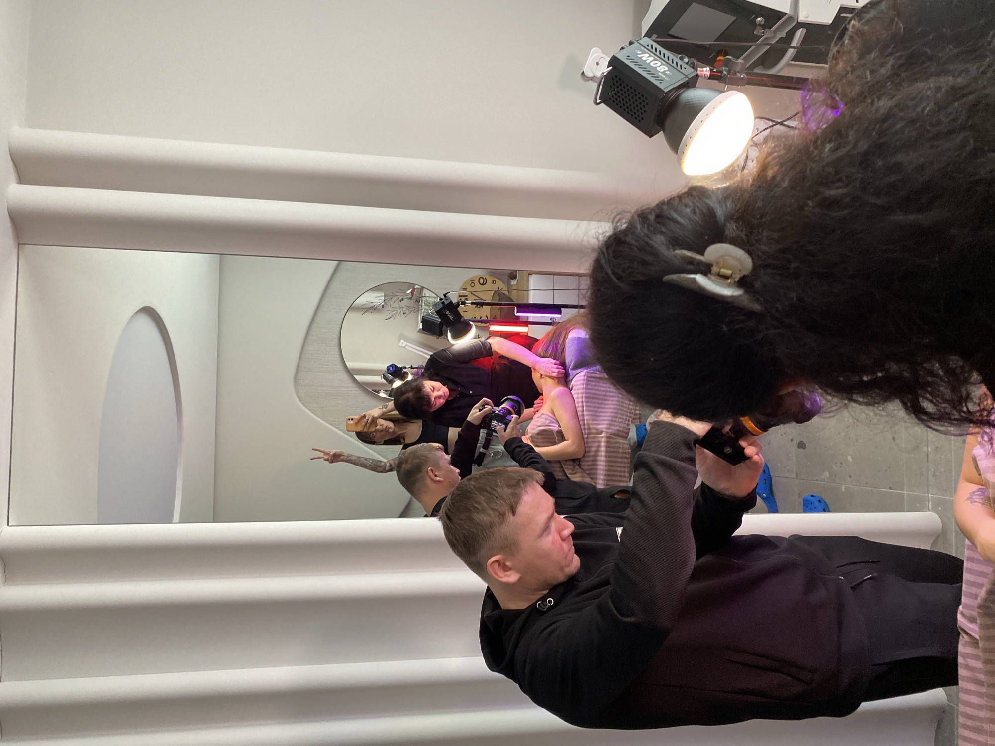 The image shows a reflective surface capturing a scene with several people. In the foreground, a man in a black hoodie is visible, holding a camera, seemingly recording the scene. In the reflection, a group of people is visible, including one person taking a photo with a smartphone, giving a peace sign. There are also individuals holding cameras and wearing various clothing. The environment appears to be a studio or a staged area with white walls and decorative elements. Overhead, there is lighting equipment. There is not enough information to determine that this photo was taken in the city of Izhevsk, Russia.