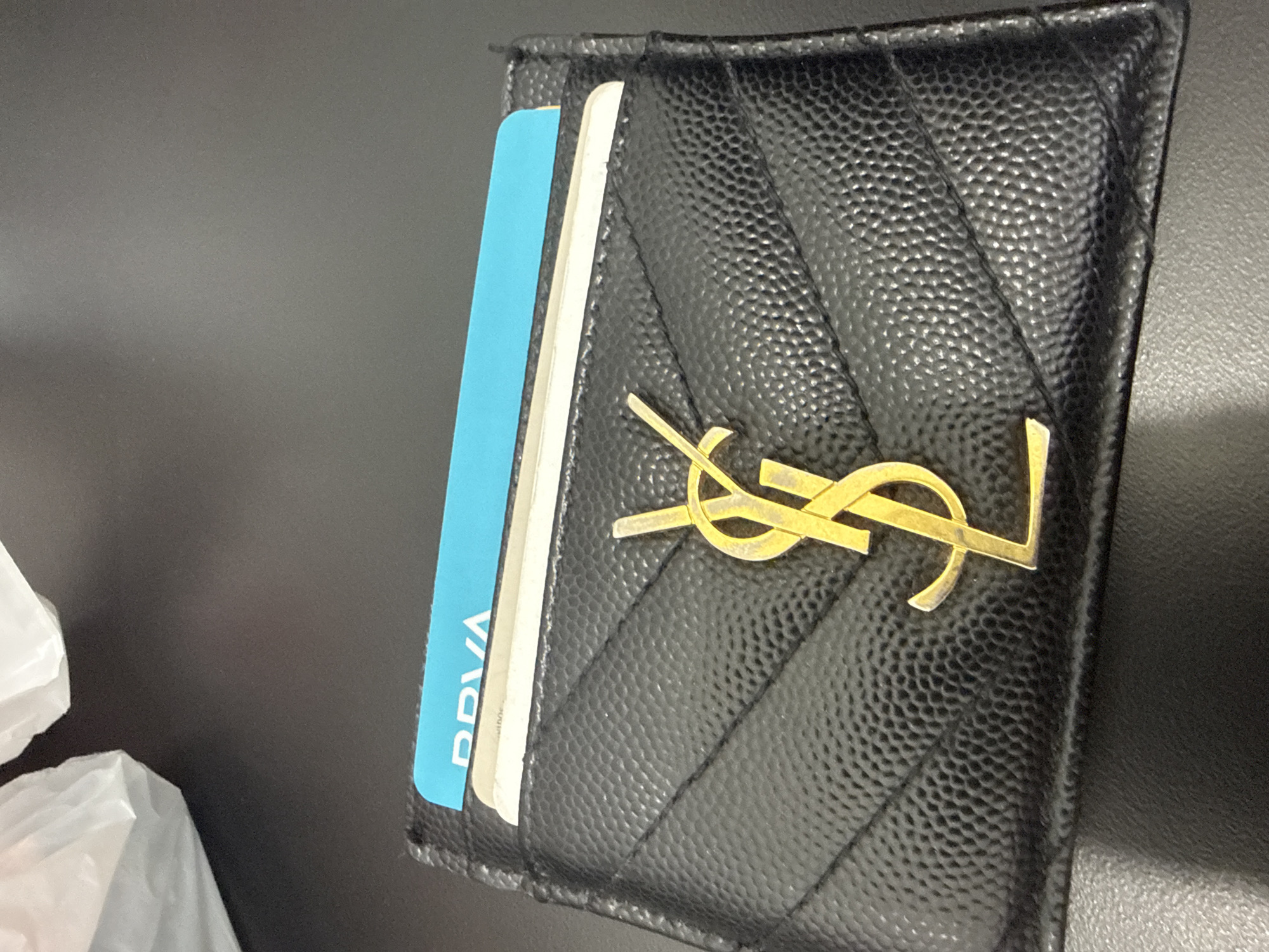 A black Yves Saint Laurent card holder with a gold YSL logo is resting on a dark surface. The card holder contains two visible cards: a blue BBVA card and a partially visible white card. A blurred white plastic bag is in the lower left corner. The location, San Bernardino Tlaxcalancingo, Mexico, is unknown and cannot be verified from the image.
