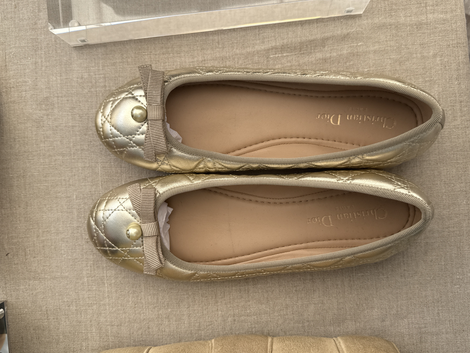 The image shows a pair of gold-colored Christian Dior ballet flats lying on a light-colored linen surface. Each shoe features a quilted pattern and a small bow detail at the toe. The interior of the shoes is a beige or tan color. The location appears to be a well-lit interior setting. There is no information present to determine if the location is in Mexico City, Mexico.