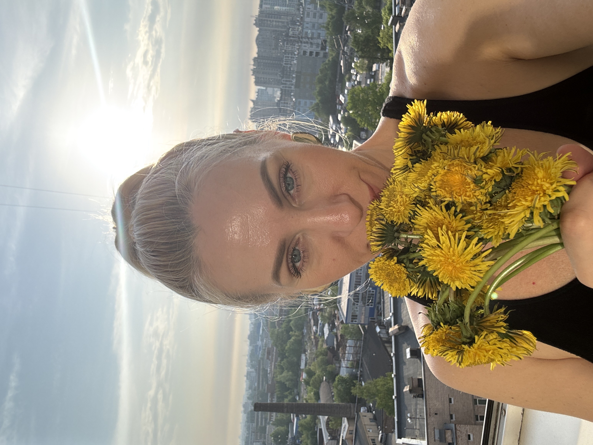 A woman with light-colored hair holds a bouquet of dandelions in front of her face. She is wearing a black tank top. The background features a city with buildings and trees. The sky is bright with the sun shining. The location appears to be an elevated position overlooking the city.