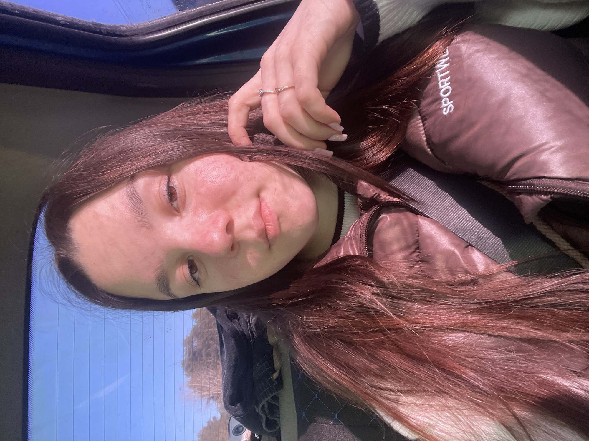 The image depicts a woman in a vehicle. The woman is wearing a brown puffer jacket with the word "SPORTWEAR" visible on the collar. She is also wearing a seatbelt. She has brown hair and is wearing rings on her fingers. The vehicle appears to be parked, as a view of a blue sky and trees is visible through the rear window. There is no specific information in the image to confirm the location as Chaikovsky, Russia.