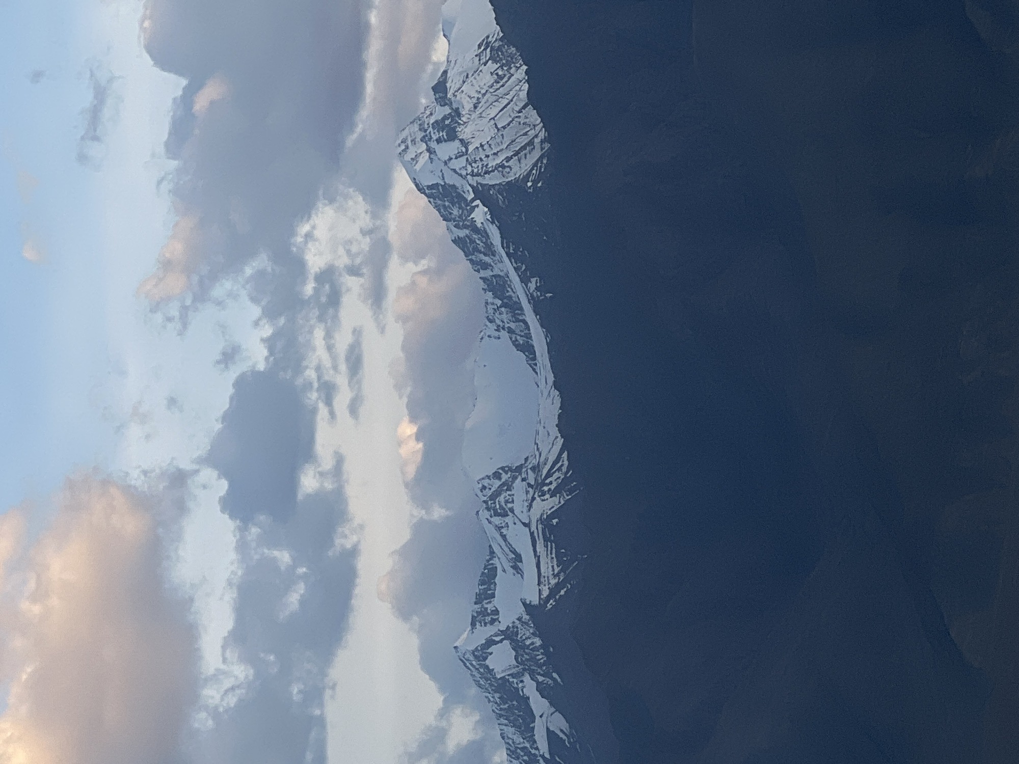 The image depicts a panoramic view of snow-capped mountains under a cloudy sky. The lower portion of the image is dominated by the dark, rugged slopes of foothills or lower mountains. Above these dark masses, the majestic, snow-covered peaks rise, intricately detailed with shadows and highlights. The sky is filled with a mix of light and dark gray clouds, with hints of pink and orange from what appears to be the setting or rising sun. The overall scene is one of natural grandeur and dramatic lighting. The location is identified as the city of Leh, India, a region known for its stunning Himalayan landscapes. There are no people or discernable man-made objects in the frame, emphasizing the natural and vast scale of the environment.