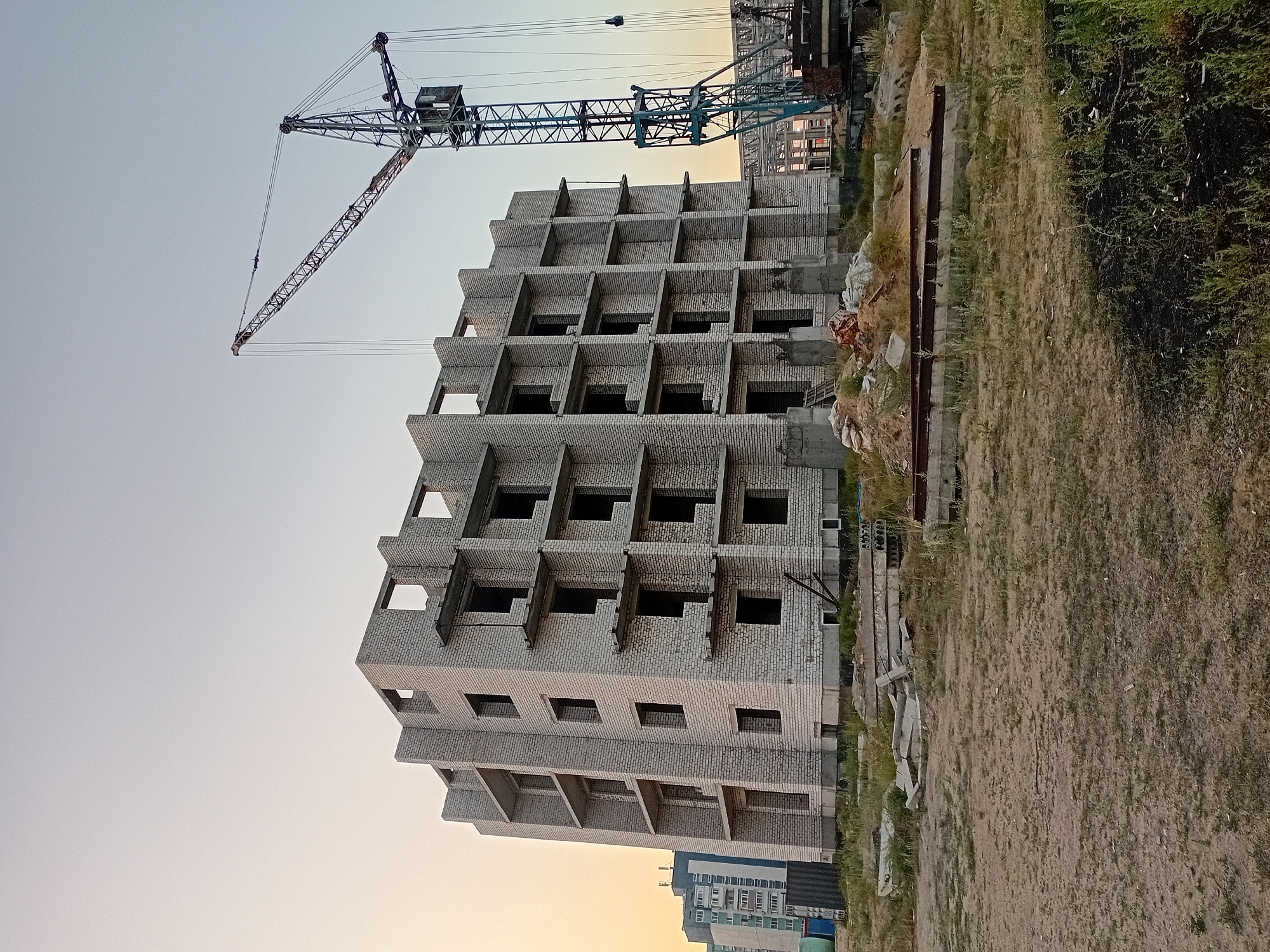 The image depicts an unfinished multi-story building under construction in Semey, Kazakhstan. The building is constructed from white bricks and has multiple floors with open window frames and unfinished balconies. A large construction crane stands to the right of the building, with its boom extended. The surrounding area is a dirt lot with scattered construction debris, including concrete slabs and piles of sand and bricks. In the background, another completed apartment building is visible, suggesting an ongoing urban development project. The sky is clear with a pale gradient from yellow at the horizon to light blue at the top, indicating either sunrise or sunset. There are no people visible in the image.