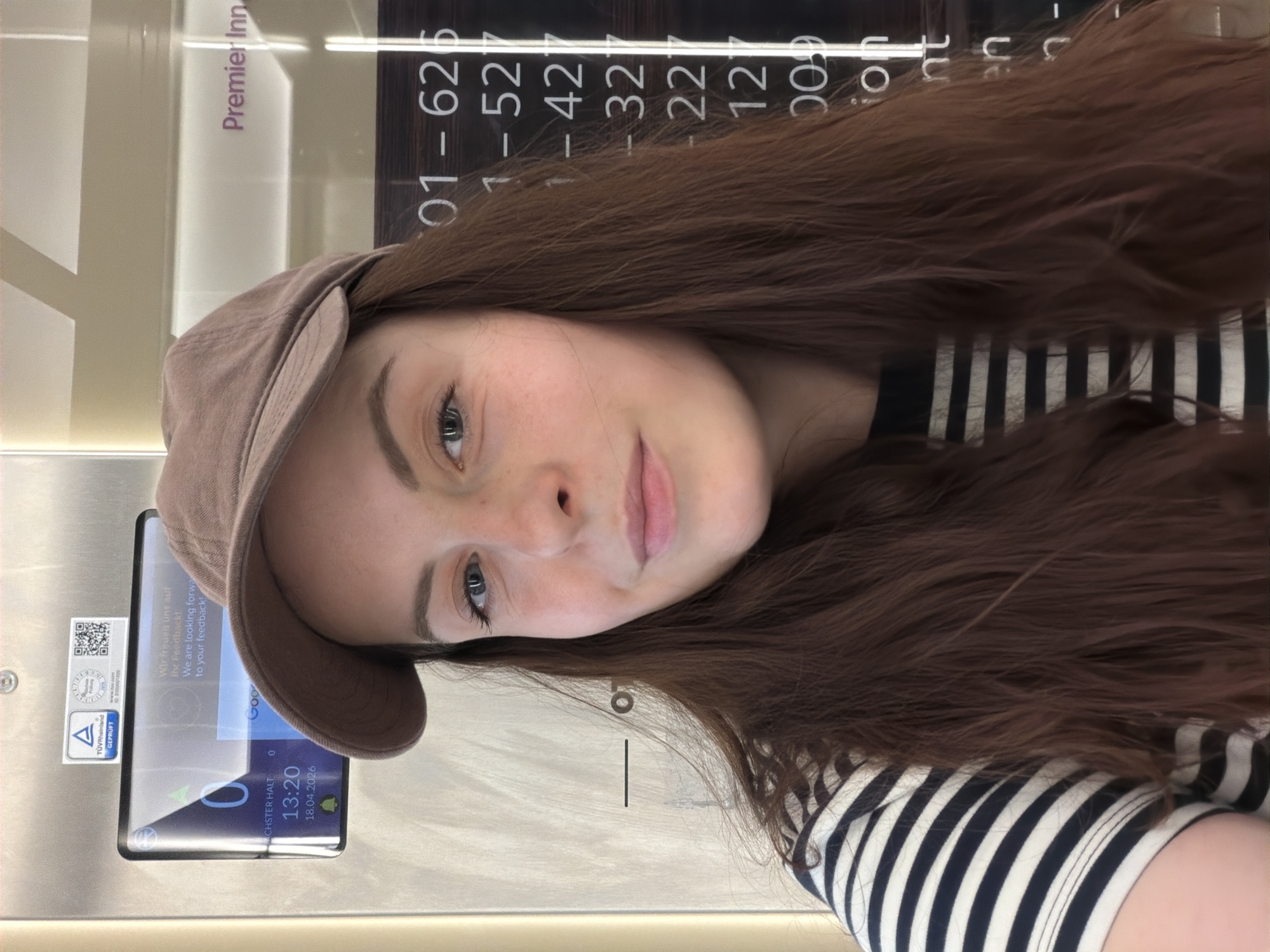 The image is a selfie taken by a young woman wearing a brown cap and a black and white striped shirt. The background includes what appears to be a ticket machine or information panel with a screen displaying the time "13:20" and the date "18.04.2026". The screen is also labeled "NÄCHSTER HALT". The panel has a sticker stating it is "TÜV Rheinland GEPRÜFT". The woman is near a hotel, specifically a Premier Inn, based on the sign partially visible. The number display "01-6251-521-421-32221200" is also visible. Based on the "NÄCHSTER HALT" and ticket machine it seems she is at a public transportation station in Wiesbaden, Germany.