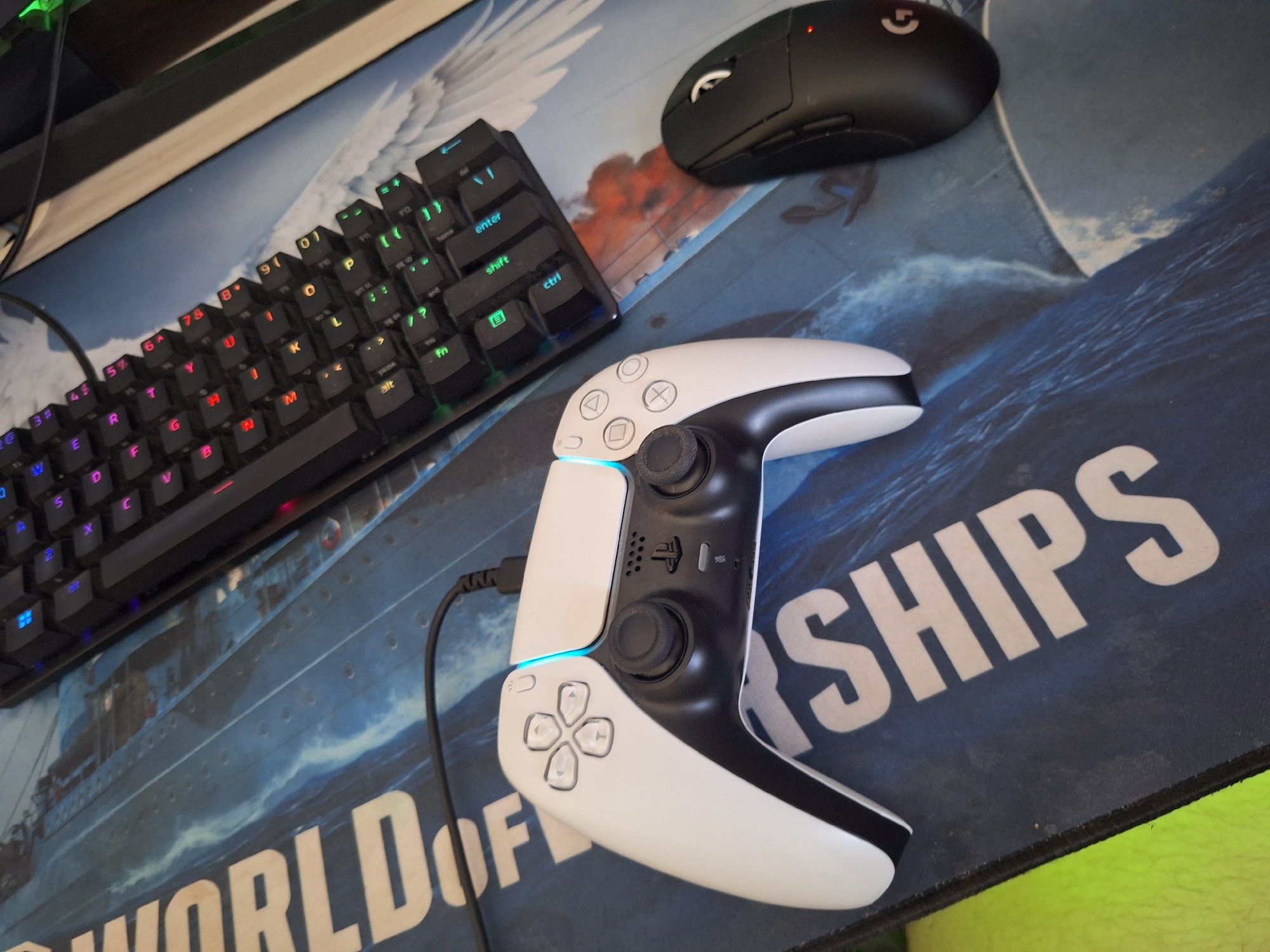 The image shows a top-down view of a gaming setup on a desk. A white and black video game controller, likely a PlayStation 5 DualSense, is positioned in the center. To the right of the controller is a black gaming mouse with a red LED indicator light and a Logitech logo. Behind the controller, a black mechanical keyboard with RGB lighting is visible. The setup is placed on a large, dark blue mousepad with a graphic design featuring the words "WORLD OF" in large white letters on the left, and "SHIPS" partially visible on the right. The background includes a portion of a desk with a light-colored surface and a dark edge. The scene appears to be indoors, likely in a home or office environment, and there is no indication of its specific location within the city of Varna, Bulgaria.