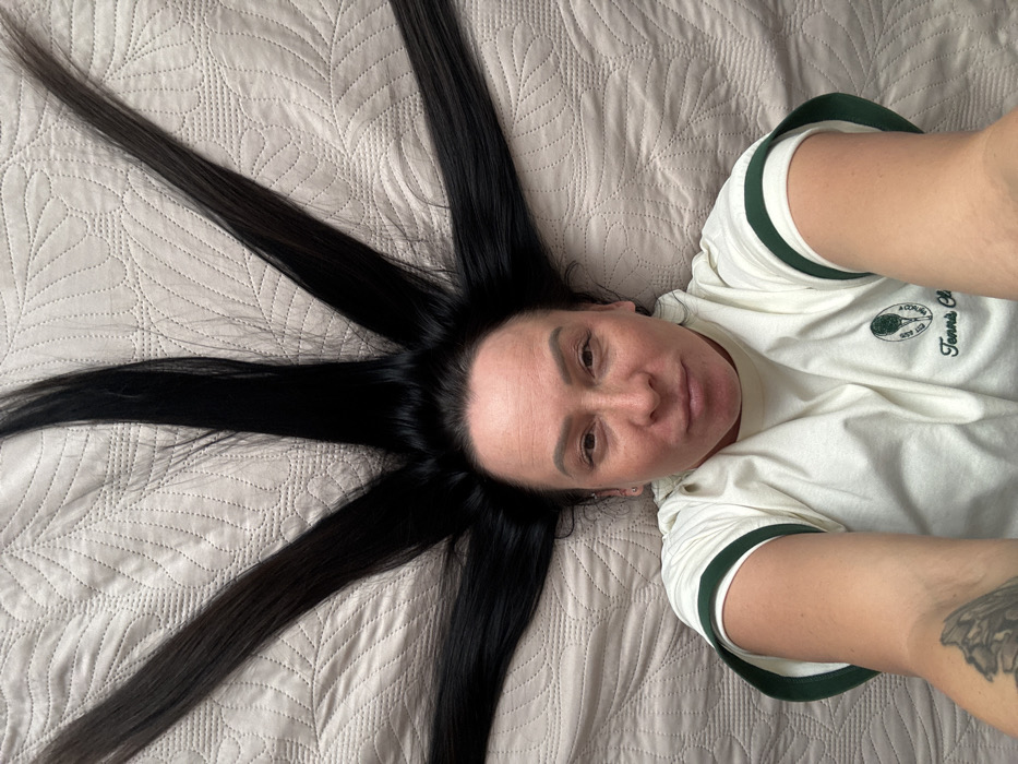 An overhead photograph captures an adult female lying supine on a light beige quilted surface, presumably a bed, within a residential interior in Pavlovsky, Russia. Her dark, straight hair is meticulously fanned out around her head, creating a symmetrical, star-like pattern against the bedding. She is dressed in an off-white or cream-colored t-shirt featuring dark green trim on the sleeves and an embroidered "Tennis Club" logo on the left chest area. Her face is directed towards the camera with a neutral expression. Her right forearm is visible, extending towards the lens, indicating she is capturing the image herself. A dark, intricate tattoo is visible on her left bicep/upper arm, partially obscured by her sleeve. The quilted surface exhibits a repetitive, stylized leaf or feather pattern.
