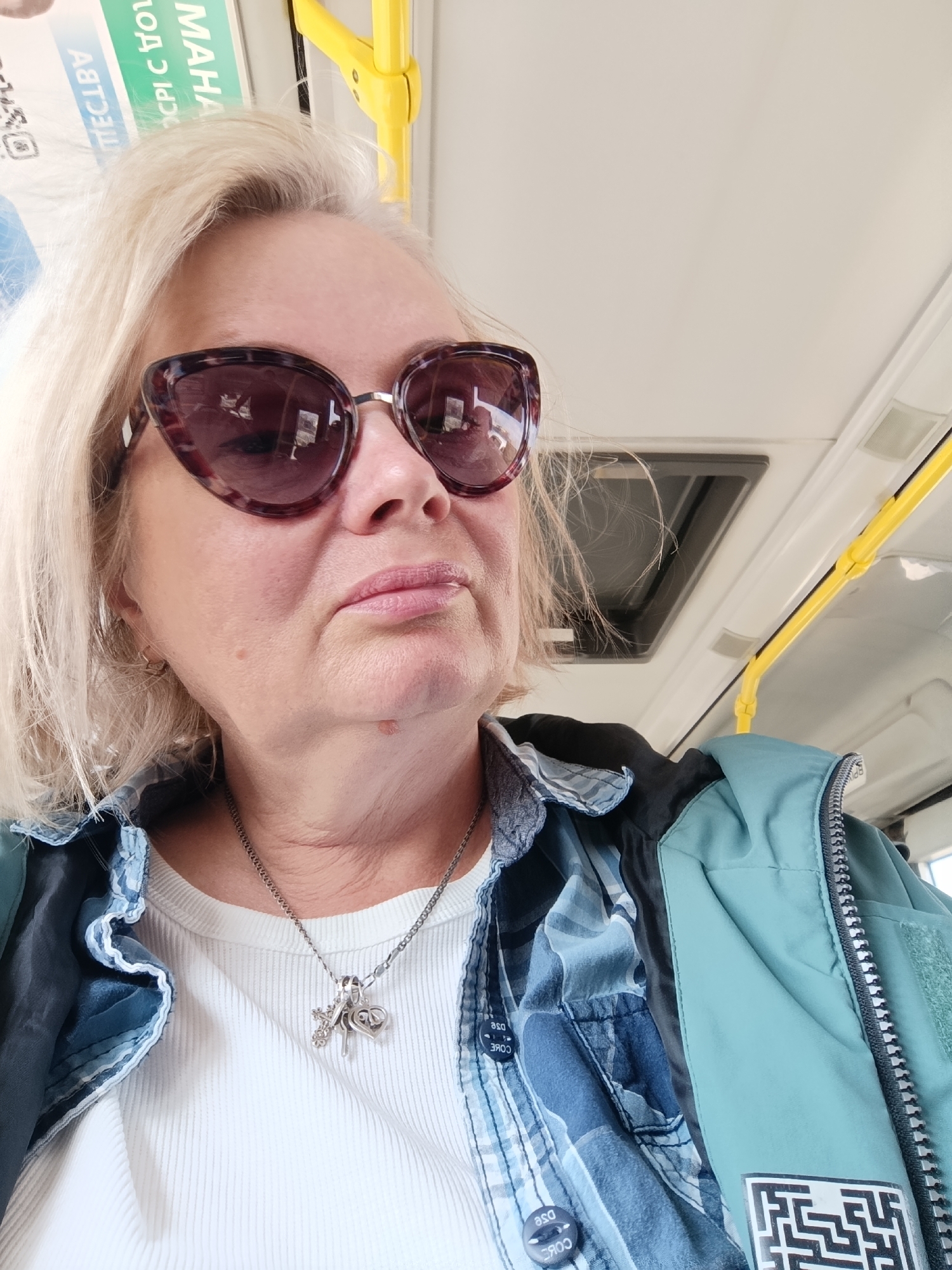 A candid, slightly low-angle shot captures a blonde woman riding in a public transport vehicle in Chaikovsky, Russia, during the daytime. The woman, who appears to be middle-aged or elderly, wears large, dark tortoiseshell-patterned sunglasses that obscure her eyes. Her expression is neutral, with lips slightly parted.
She is dressed in layers, consisting of a white ribbed long-sleeved top, a blue and white plaid shirt with "CORE DSE" visible on its buttons, and a teal or mint green zippered jacket. A silver necklace with multiple charms, including a cross, a key, and a heart inscribed with "love," is visible around her neck. Her blonde hair is loosely styled and frames her face.
The background elements confirm the setting as a public vehicle, likely a bus or tram. A bright yellow grab rail is prominent on the right side of the frame, and an overhead vent or light fixture is visible. In the upper left, a partially obscured and mirrored advertisement or sign shows Cyrillic text, including fragments like "МАЙНА" and "с ОРСК," along with mirrored letters such as "АЯТЯО" and "ТЕШСВ". A black and white maze-like pattern is visible on a patch on her jacket. The lighting is bright, suggesting a clear day. The woman is looking towards her right, off-camera.