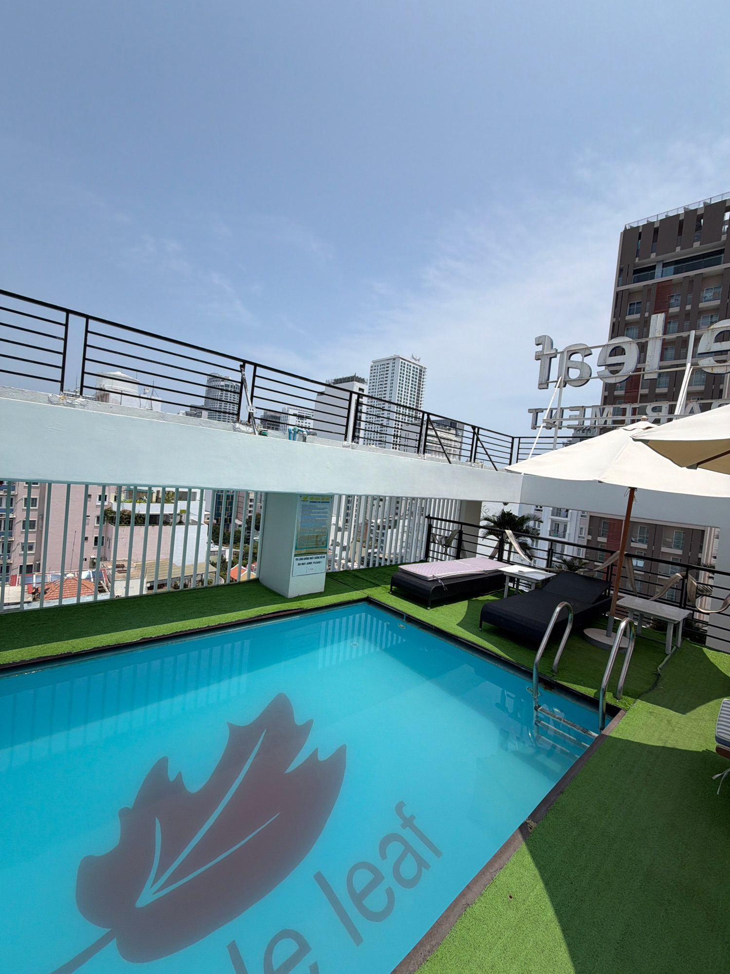 The image shows a rooftop swimming pool with a blue leaf logo at the bottom, located in Nha Trang, Vietnam. The pool is surrounded by green artificial turf, and there are lounge chairs, a table, and umbrellas for relaxation. In the background, tall buildings and the sky are visible. A sign with text in Vietnamese is mounted on a white pillar near the pool. The overall scene depicts a resort-style amenity in an urban setting.