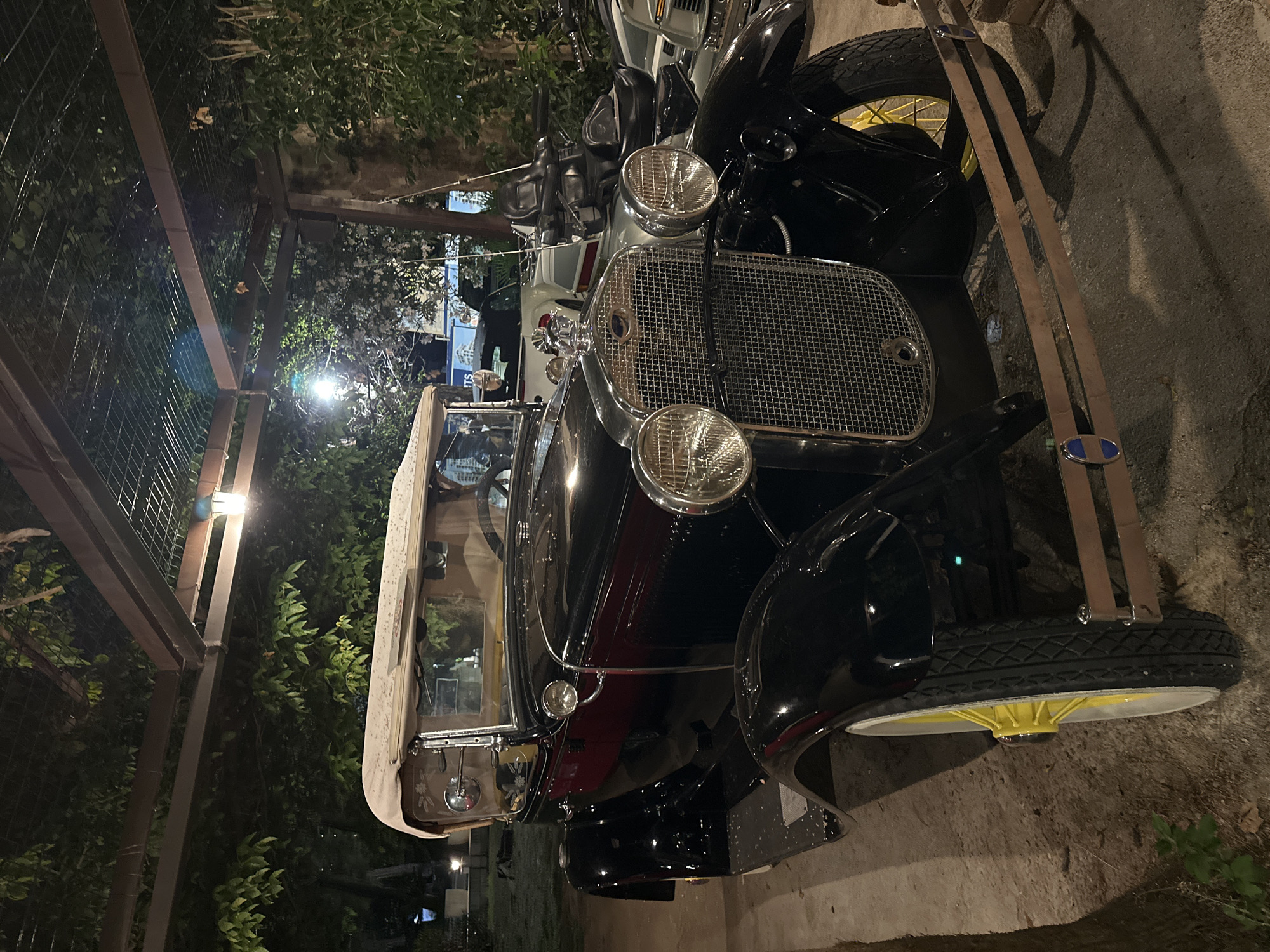 The image captures a vintage black convertible automobile parked outdoors, likely in Budva, Montenegro. The car is the primary subject, featuring a cream-colored retractable roof, chrome grille, and two large headlights. The wheels have white rims with bright yellow spokes. The car appears to be in good condition, suggesting it is either restored or well-maintained.
In the background, other vehicles are visible, including a motorcycle with a large passenger seat and what appears to be another car. The setting is an outdoor area, possibly a courtyard or parking lot, with foliage and trees surrounding the vehicles. A pergola-like structure with wooden beams and a wire mesh ceiling is overhead, with lights illuminating the scene. The ground is a mix of concrete and dirt. The lighting suggests it is either evening or night. No people are clearly visible in the frame.