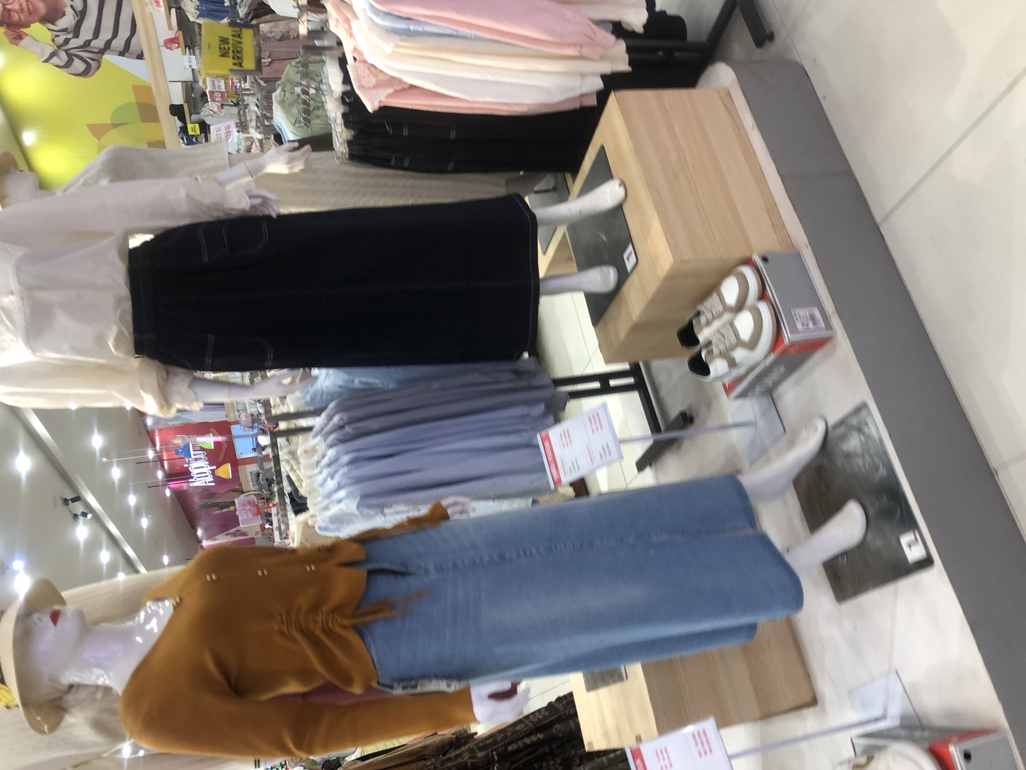 The image depicts a retail store interior, likely a clothing boutique, with mannequins dressed in casual wear. One mannequin on the left is adorned with a rust-colored, ruched long-sleeved sweater and a light blue denim maxi skirt, accessorized with a straw hat. Another mannequin to the right wears a white long-sleeved blouse and a black denim maxi skirt. In the foreground, a pair of white and tan sneakers are displayed on a box. Racks of clothing, including light blue shirts and pastel-colored blouses, are visible in the background, along with store signage and lighting fixtures. The store appears to be organized and well-lit, typical of a commercial establishment. No specific details about Langsa, Indonesia, are discernible from the image itself.