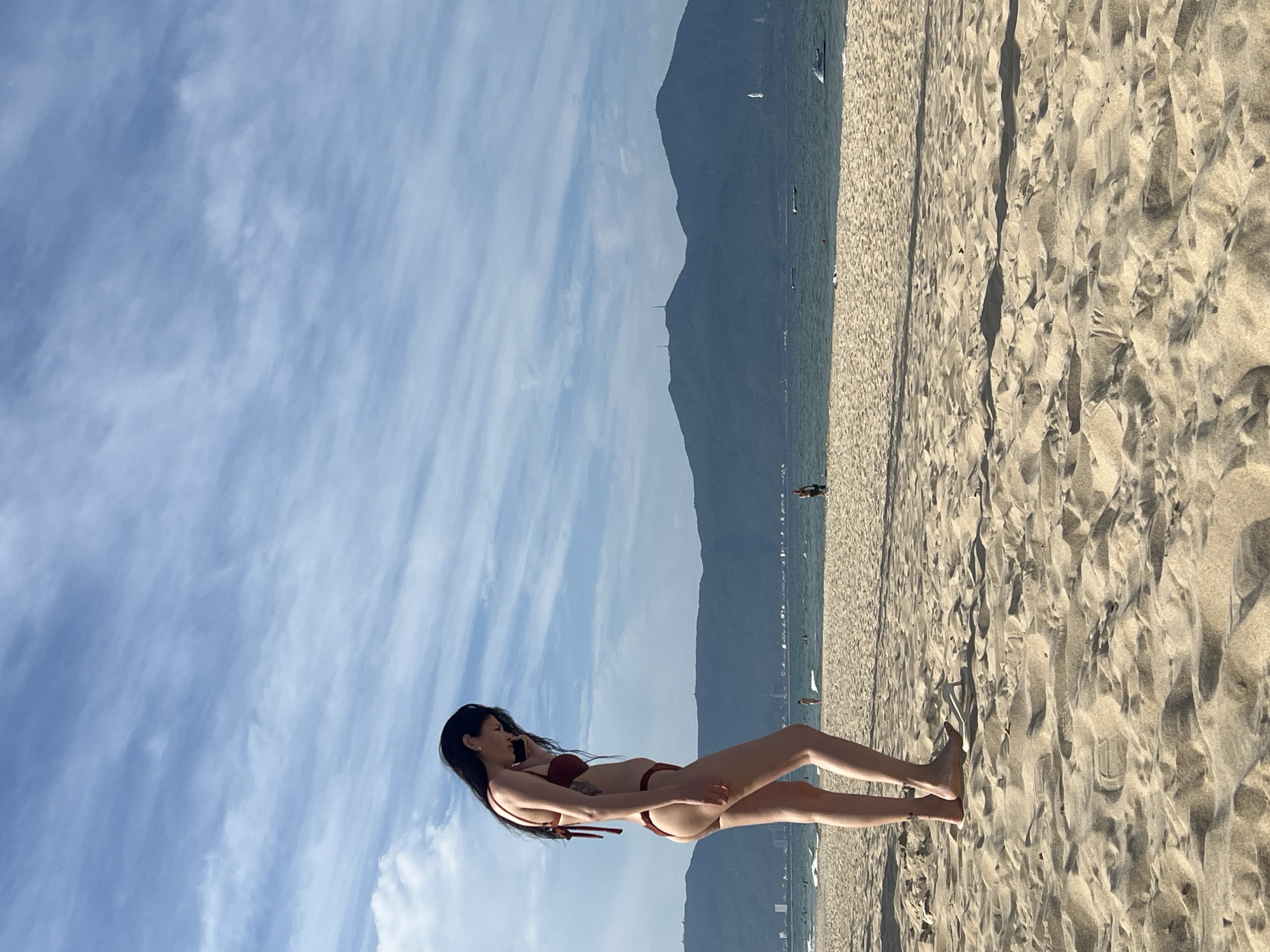 A woman in a maroon bikini stands on a sandy beach, looking down. She has long black hair and a tattoo on her left side. The beach is wide and covered in sand, with footprints visible. In the background, the turquoise ocean meets a hazy shoreline with mountains and buildings. Several small boats and a jet ski are scattered on the water. The sky is blue with wispy white clouds. The scene is set in Đà Nẵng, Vietnam, likely at My Khe Beach.