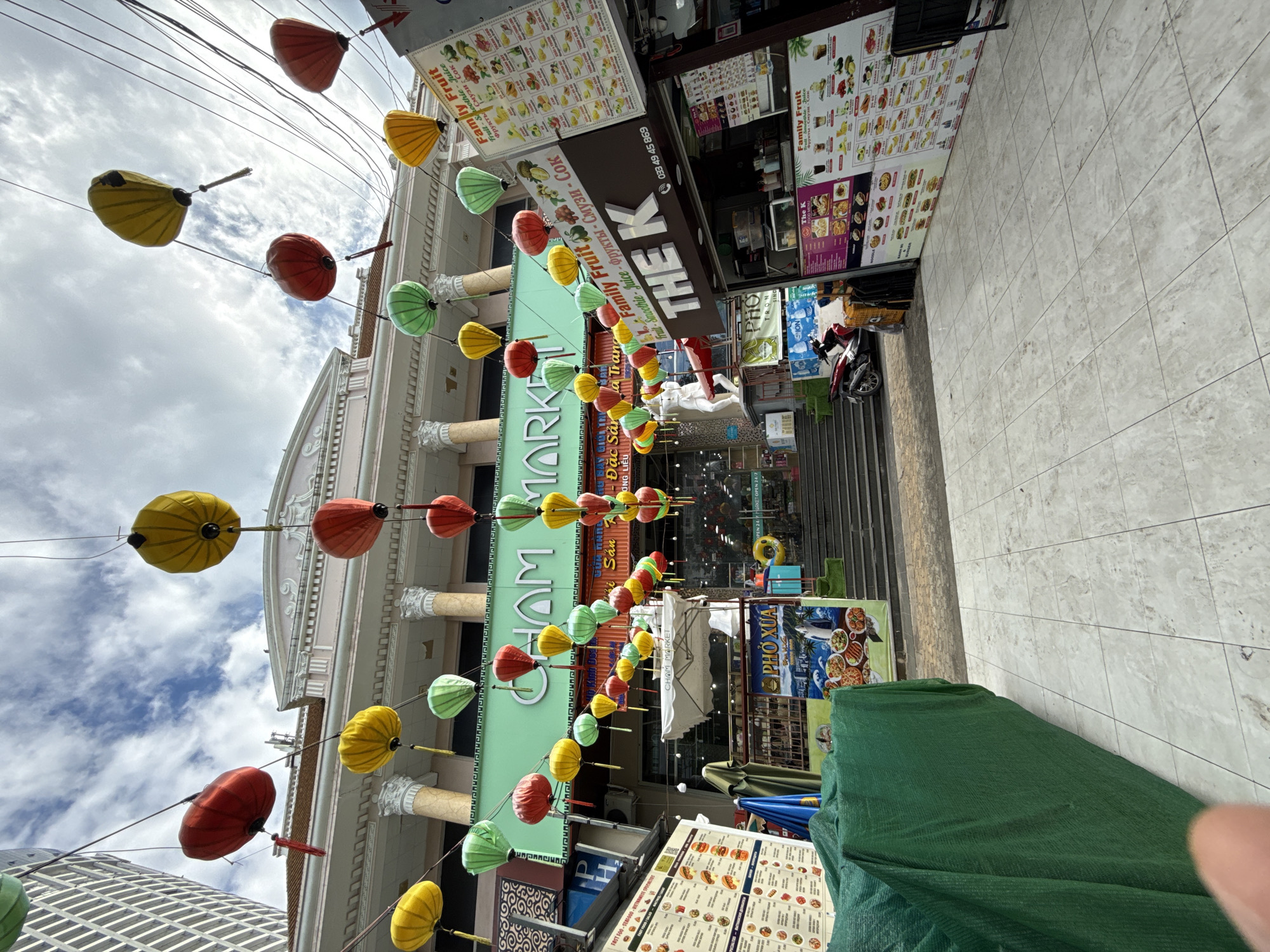 The image depicts a street scene in Nha Trang, Vietnam, characterized by a vibrant array of colorful lanterns strung across the street. In the background, a multi-story building with classical architectural elements, including columns and ornate detailing, serves as the backdrop. The lower portion of this building features signage for "CHAM MARKET," with the name displayed prominently in large green letters.
Below the market entrance, a series of steps leads down to the market itself, with various stalls and shopfronts visible. To the right of the steps, a stall named "THE K" displays a menu for "Family Fruit" with listings for fruits, smoothies, and juices in English and Russian. Further to the right, a sign advertises "Family Fruit" with a colorful selection of fruits. A red scooter is parked on the left side of the steps.
In the foreground, a large green tarp covers an area, suggesting construction or storage. The ground is paved with light-colored square tiles. The sky above is partly cloudy. No people are clearly visible in the scene.