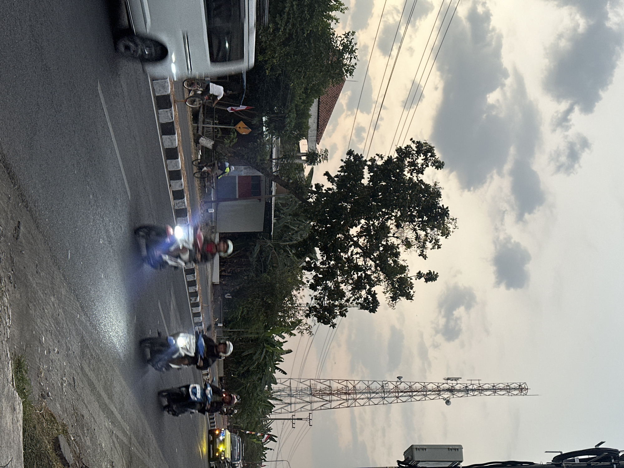 This image captures a street scene in Bulakrejo, Indonesia, during what appears to be dusk or early evening. The sky is overcast with scattered clouds, casting a diffused light over the environment. A prominent feature is a tall, lattice radio tower situated behind a cluster of trees.
In the foreground, a paved road is visible with a white line marking the lanes and a raised curb with black and white stripes. Several vehicles are present on the road, including a white van on the left side, its motion suggested by a blur. Three motorcycles, each with a rider, are moving towards the viewer from the right. The headlights of the motorcycles are illuminated, casting beams onto the road surface. A yellow vehicle is also visible further down the road on the right.
On the side of the road, a few individuals on bicycles can be seen. One person is riding a bicycle near a small building with a red and blue facade. Another individual is also on a bicycle further down. Vegetation, including trees and banana plants, lines the roadside, partially obscuring buildings in the background. Power lines are strung across the sky, supported by utility poles. The overall impression is of a suburban or semi-rural area experiencing the transition from day to night.