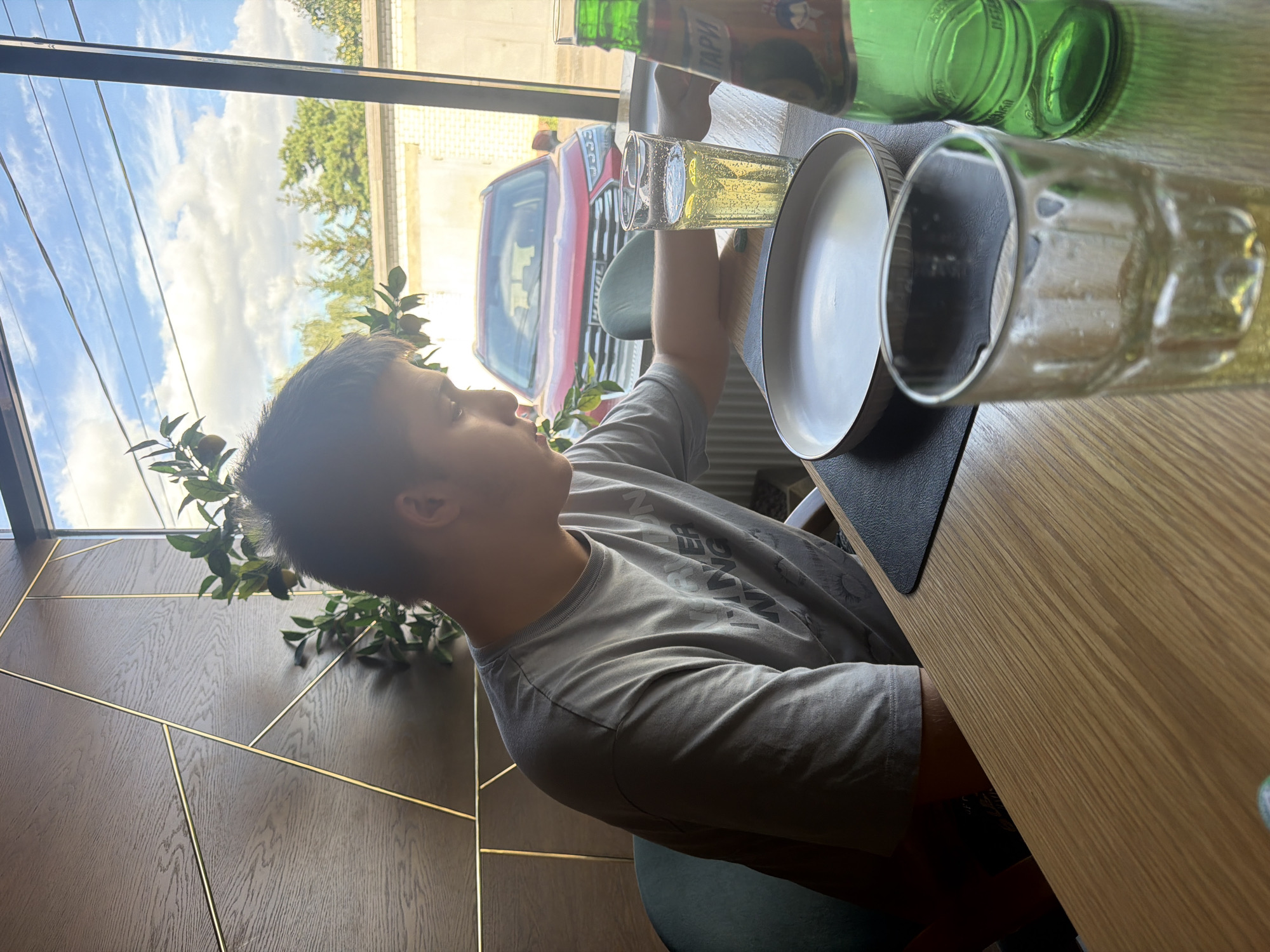 A young male with short dark hair is seated at a wooden table in a cafe. He is wearing a grey t-shirt with a graphic print. In front of him are two glasses, one with a light-colored beverage and ice, and another empty glass. A white plate and a green glass bottle are also on the table. The cafe features a modern interior with a dark wood paneled wall adorned with gold geometric accents. Behind the male, through a large window, a red SUV is parked outside. The sky is cloudy. The cafe appears to be located in Kazan, Russia, suggested by the bottle label which reads "Тархун" (Tarkhun), a popular Russian soft drink.