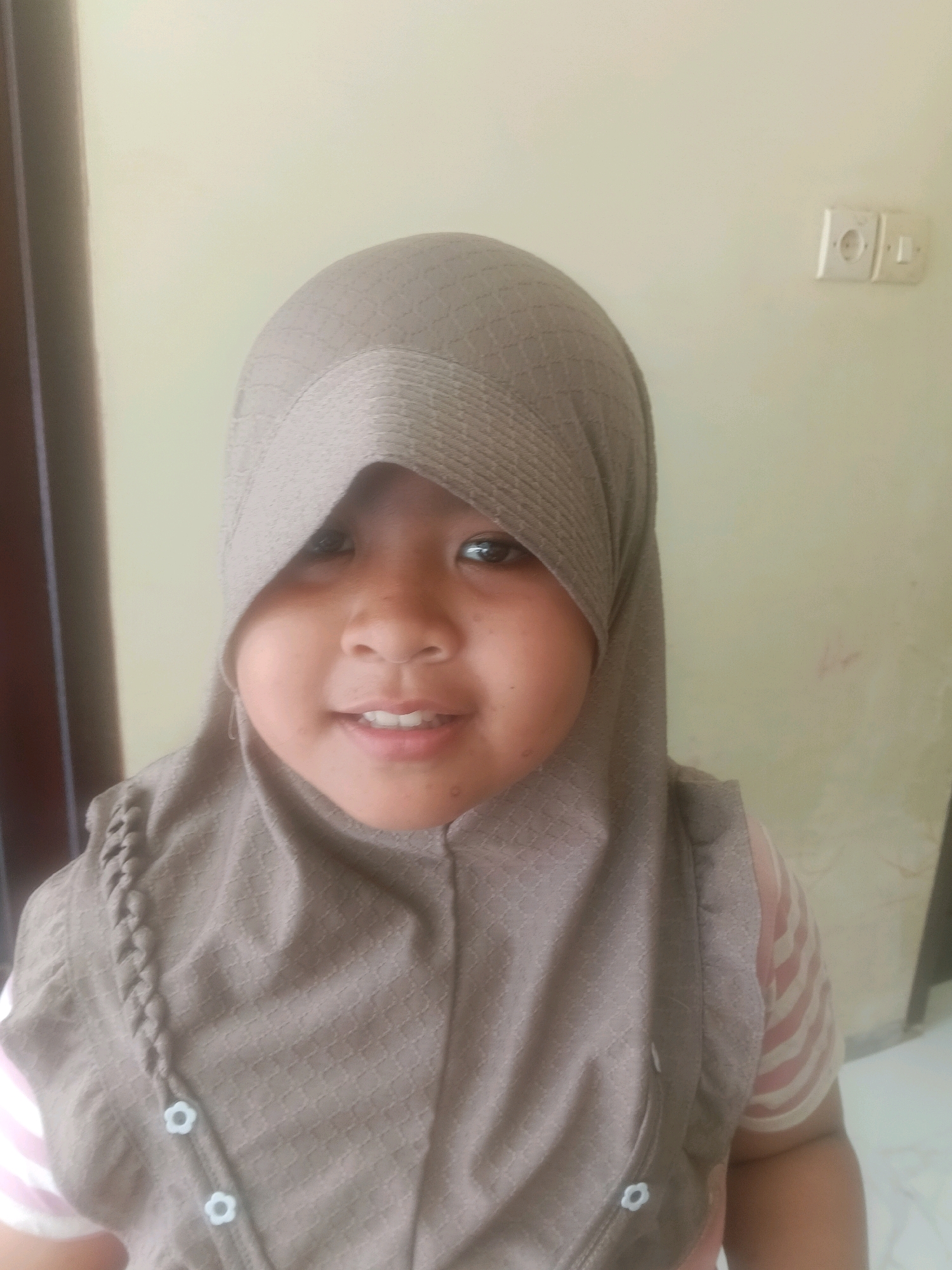 A young girl, likely of Indonesian ethnicity, is pictured in a close-up shot. She is wearing a light brown hijab that drapes over her head and shoulders, partially obscuring her forehead. Underneath the hijab, her face is visible, with dark eyes, a small nose, and a smiling mouth revealing straight, white teeth. Her skin appears to have a light tan complexion. She is also wearing a brown vest with a textured pattern, adorned with small white flower-shaped buttons down the front. A braid detail is visible on the left side of the vest. A hint of a striped pink and white garment can be seen beneath the vest on her right arm. The background is a plain, light-colored wall, with an electrical outlet visible on the right side of the frame. The overall lighting suggests an indoor setting, possibly a home in Bulangan Barat, Indonesia, given the typical architectural features like the wall and electrical outlet.
