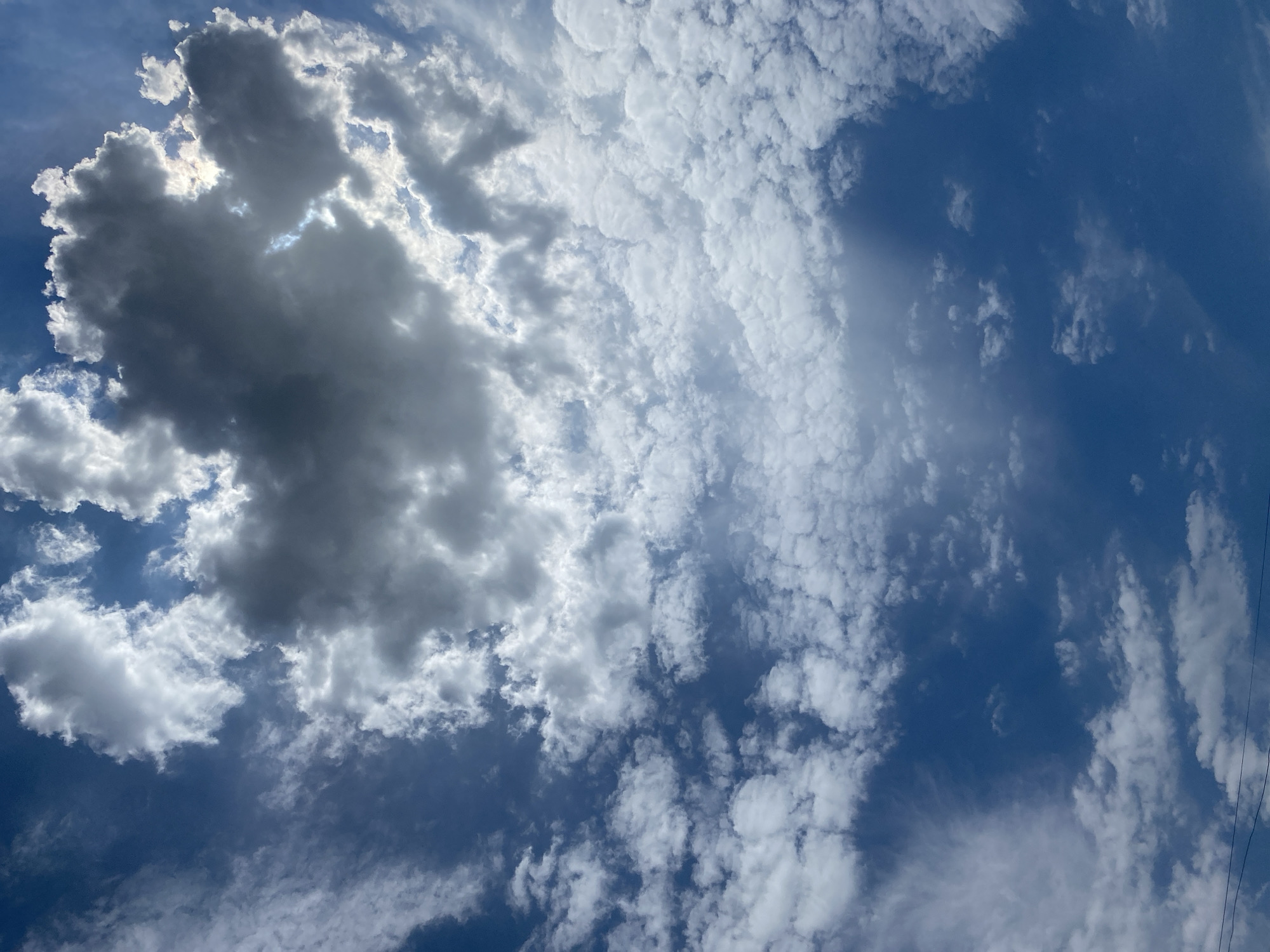 The image shows a bright blue sky filled with a variety of clouds. In the center, a large, dark, and dense cloud formation dominates the frame, partially obscuring the sun, which creates a bright halo around its edges and casts rays of light through breaks in the clouds. To the left and right of this central mass, smaller, scattered white cumulus clouds drift across the sky. Further down, the clouds become more wispy and delicate, appearing as thin streaks against the deep blue. The bottom of the image shows a sliver of a dark horizon line, possibly indicative of distant land or structures, and two thin, dark horizontal lines that could be power lines or wires. The overall impression is of a dynamic sky with contrasting cloud types and dramatic lighting. The image was captured in Gurlan, Uzbekistan.
