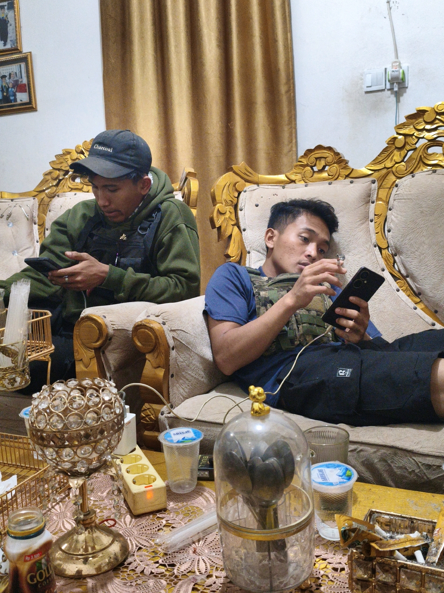 In a dimly lit room, two individuals are seated on a plush, ornate couch. The room appears to be a living area, with golden curtains and a white wall. On the left, a person wearing a green hooded sweatshirt and a tactical vest is looking down at their phone, which they are holding in their left hand. A black baseball cap with "Charcoal" printed on it is on their head. A ring is visible on their left ring finger.
To the right, another person, also wearing a tactical vest and a blue t-shirt, is looking at their phone, which they are holding in their right hand. They are wearing black shorts. A charging cable is plugged into their phone and runs towards the floor.
On a table in front of the couch, several objects are visible. These include a decorative crystal sphere on a gold stand, a power strip with an orange illuminated outlet, a plastic cup with a blue lid, a bottle of "Gold Coffee," a glass of water, and an ashtray overflowing with cigarette butts. The table is covered with a patterned lace tablecloth.
The overall impression is one of a relaxed, indoor setting, with the two individuals engaged with their mobile devices. The presence of tactical vests on both individuals suggests a potential professional context, possibly law enforcement or security personnel, though this cannot be definitively determined from the image alone. The location is identified as Koto Baru Semurup, Indonesia.