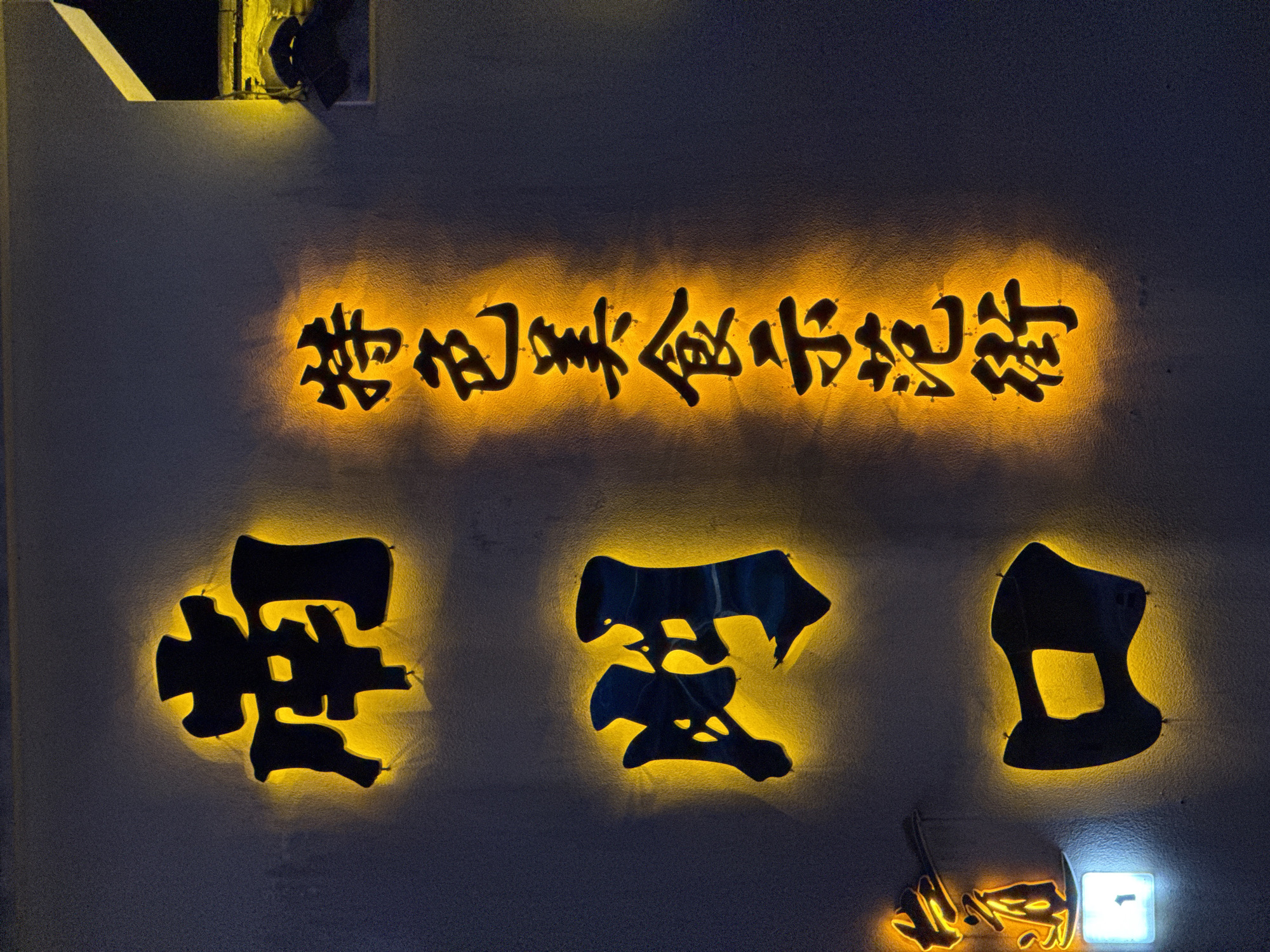 The image depicts a sign on a wall in Zhangjiajie, China. The text is in Chinese characters, illuminated from behind with a warm, yellow light. The top line of text reads "Zhangjiajie Meishi Xiaochi" (張家界美食小吃). Below this, three larger characters are arranged in a row. To the left of the frame, there is a light fixture on the upper left side. Another light fixture with a brighter, white light is visible towards the lower right of the frame. The wall has a slightly rough texture.