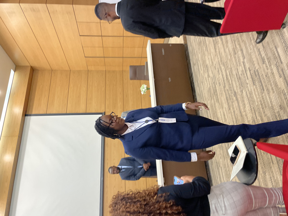 The image depicts an indoor scene from what appears to be a formal conference, meeting, or presentation in Luanda, Angola. The primary subject is a woman with dark dreadlocks and glasses, dressed professionally in a navy blue pantsuit over a white shirt. She is standing upright, looking towards the left side of the frame, and wears a blue lanyard with an ID badge that prominently features the word "AFRICA" and a blue and white logo, suggesting participation in an African forum or event.
Behind her, another man in a dark suit stands facing away, partially visible behind a light-colored table or podium adorned with a small white floral arrangement. To the left, a large white projection screen is mounted on the wall, indicating that a presentation or visual content is part of the event. A third man in a dark suit is visible in the distant background, further to the left, behind the main standing woman.
In the foreground, the back of a person's head with curly hair is visible, holding a smartphone. A small black stand with a laptop or tablet rests near this person. The venue features light-colored wooden paneling on the walls and a textured, patterned carpeted floor. Red chair backs are discernible on the right side of the frame, hinting at an audience area. The overall setting is professional and formal, illuminated by indoor lighting.