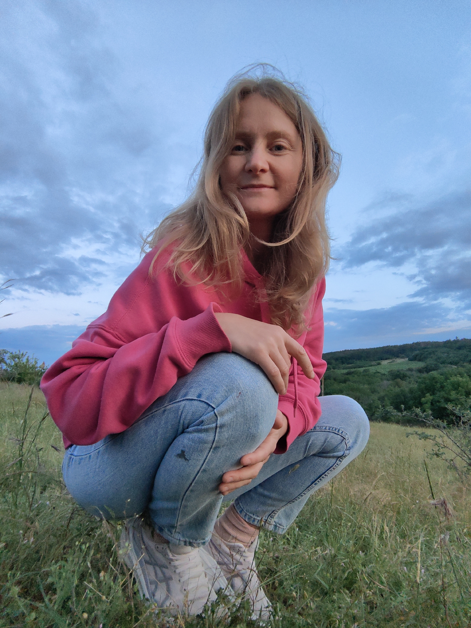 A young woman with blonde hair is crouching in a grassy field with a rolling, tree-covered landscape in the background. She is wearing a pink hooded sweatshirt and light blue jeans. The sky above is cloudy and appears to be either early morning or late evening. The overall mood is casual and relaxed. The image is taken from a low angle, with the woman looking directly at the camera.