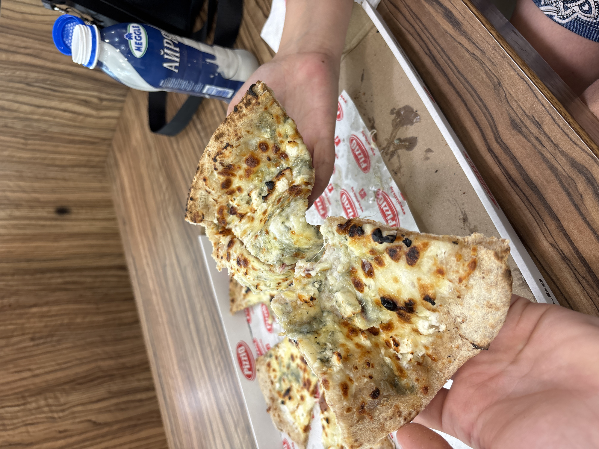 Two hands are holding slices of pizza over a pizza box. The pizza appears to be a four-cheese pizza, with visible chunks of white and blue cheese. The crust is golden brown with some charred spots. The pizza box has a logo that says "Pizzo". To the right of the pizza box, a bottle of "Meggle" brand yogurt drink is visible, with Cyrillic lettering on the label. The setting appears to be a restaurant or eatery, with a wooden table and a wood-paneled wall in the background. The overall scene suggests a casual meal. The specific location in Stara Zagora, Bulgaria, cannot be determined from the image alone.