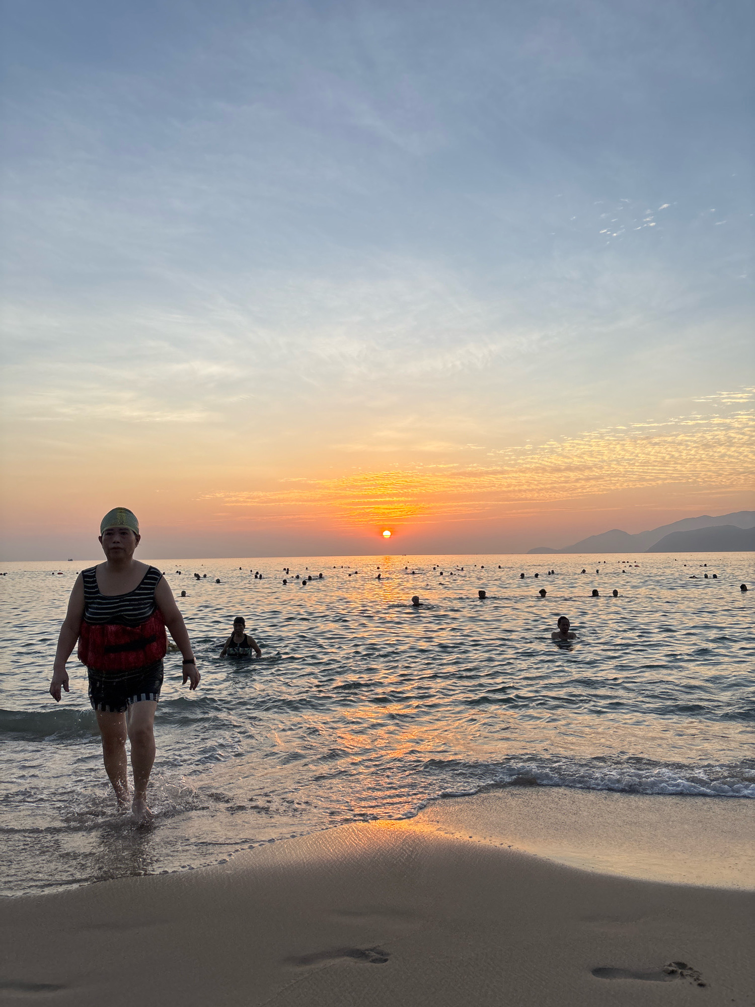 A wide-angle, eye-level outdoor shot captures a sunset scene on a beach in Nha Trang, Vietnam. The sun, a bright orange orb, is positioned just above the horizon line in the center of the frame, casting a warm glow across the sky and water. The sky transitions from a soft blue at the top to shades of orange and yellow near the horizon, with wispy clouds scattered across the upper half.
In the foreground, a person is walking out of the water onto the sandy shore. They are wearing a green swim cap, a black and white striped tank top with a red life vest over it, and black and white striped shorts. Their feet are bare and submerged in the shallow water, with small waves lapping at their ankles. The wet sand shows footprints.
In the middle ground, the ocean is filled with numerous people swimming and wading. They are mostly visible from the shoulders up, enjoying the water. The water is calm with gentle ripples.
The background features a hazy, mountainous coastline on the right side of the frame. The overall atmosphere is peaceful and serene, indicating a leisurely evening at the beach.