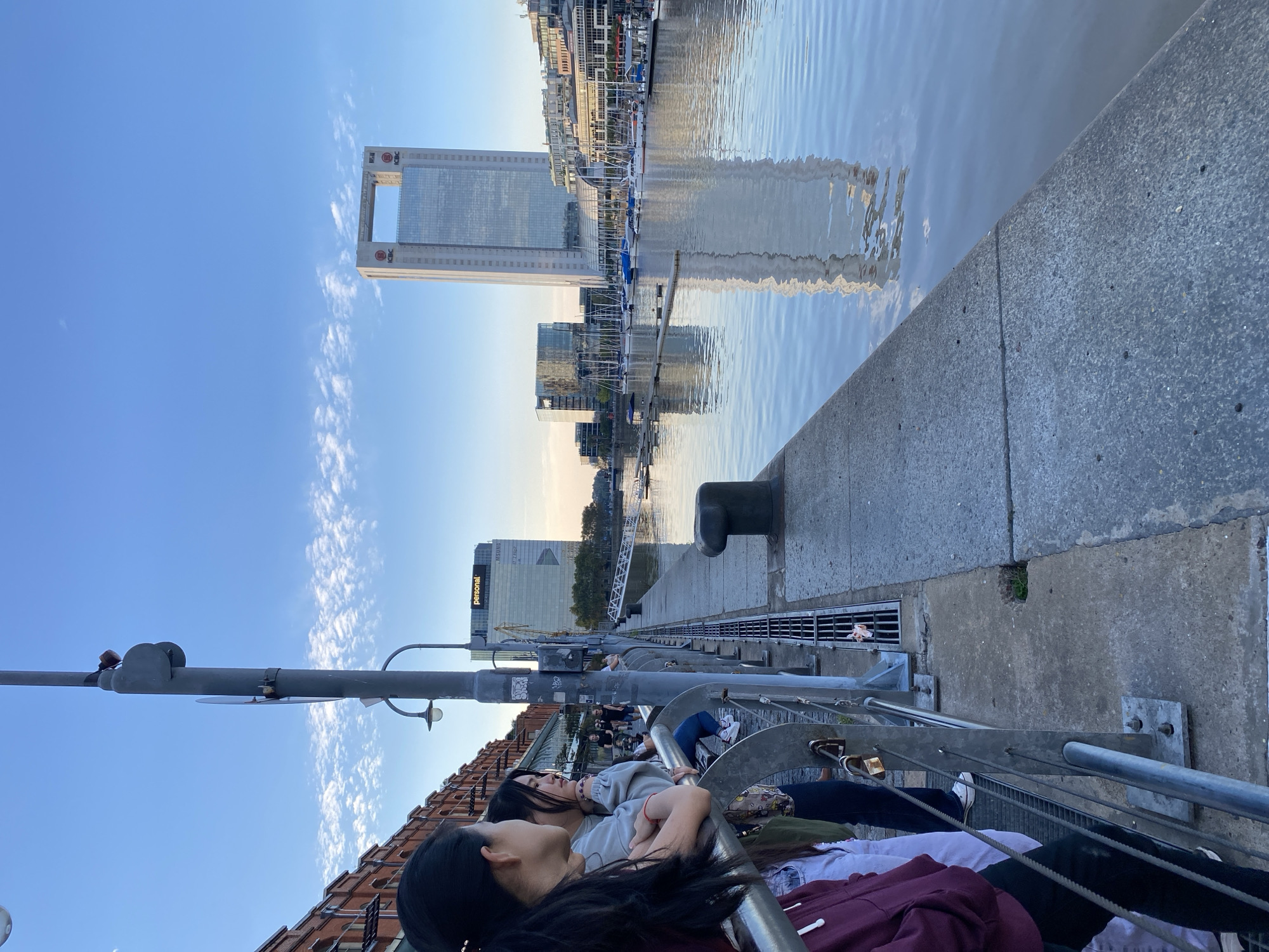 The image depicts a waterfront scene in Buenos Aires, Argentina, likely in the Puerto Madero district.
People: Several individuals are present, lounging behind a metal railing along the waterfront. Two women are closest to the camera.
Subjects/Objects: Key objects include the high-rise building with the distinctive "arch" at the top, a landmark in Puerto Madero. The scene also features water, a concrete embankment/pier, a black bollard, a lamp post, and other modern buildings.
Environment: The setting is along a waterway with calm water reflecting the buildings and sky. The sky is a clear blue with some thin, wispy clouds. The perspective of the photo suggests a waterfront promenade or walkway.