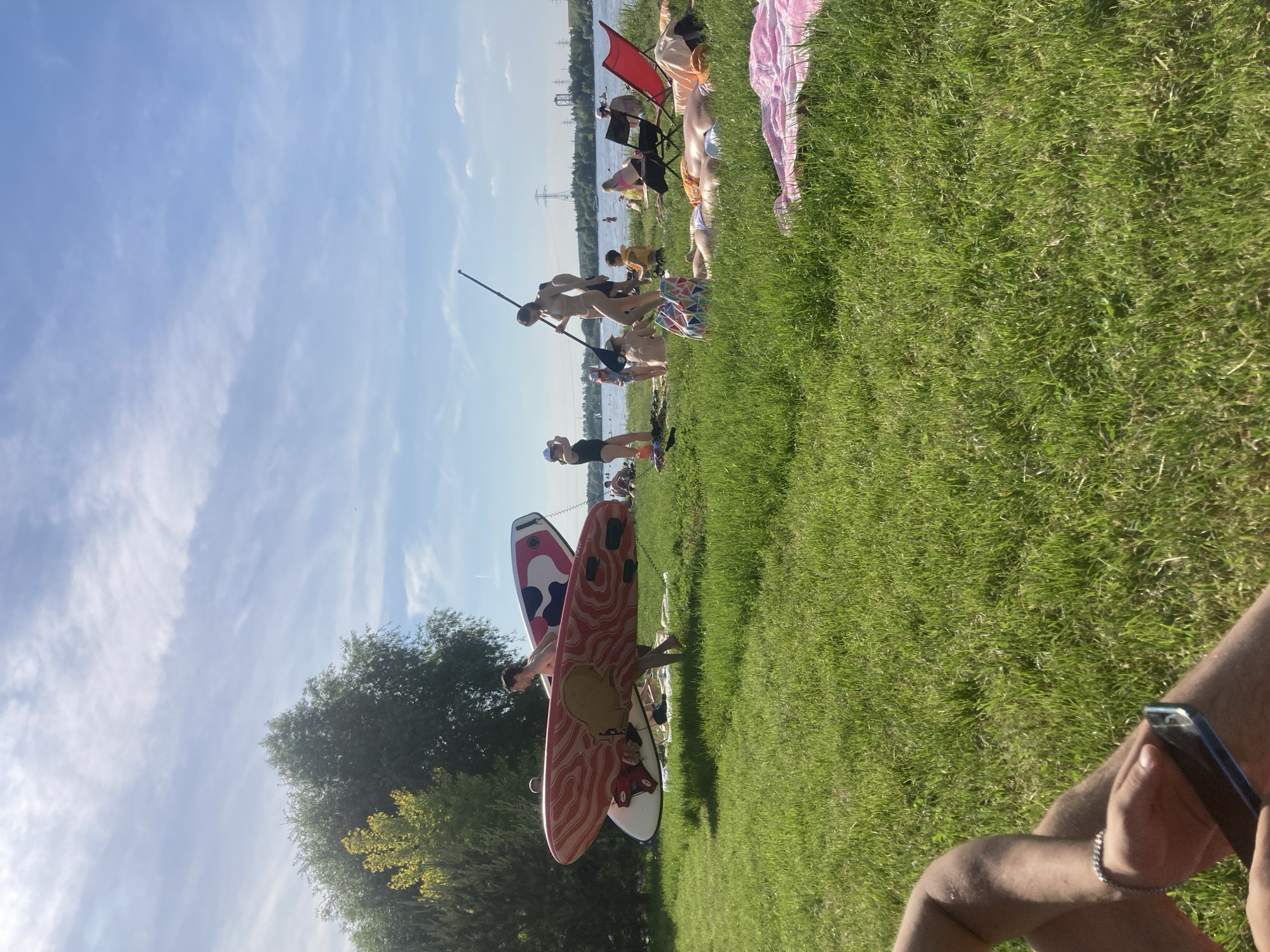 This image captures an outdoor recreational scene on a sunny day in Kapustino, Russia, adjacent to a large body of water. The foreground and mid-ground are covered in lush, vibrant green grass. The sky is bright blue with scattered, wispy white clouds.
In the bottom left corner, partially visible, are the legs and arms of an individual holding a dark-colored smartphone; a silver-toned bracelet is present on the left wrist.
Mid-ground on the left, a person, barefoot, carries two stacked stand-up paddleboards. The uppermost board is predominantly red with a swirling white pattern and a central, brown, stylized octopus-like design. The lower board features a white base with pink and black abstract patterns. Behind this individual, a portion of another person's head is visible. To the far left, dense green trees are present.
Towards the center and right mid-ground, closer to the water, a woman in a light-colored, possibly beige, swimsuit stands holding a black paddle. Another individual in a similar swimsuit is partially obscured behind her. A multi-colored beach bag with a geometric pattern rests on the grass nearby. Various people are distributed across the grassy area, some sitting or lying on towels; one person lies prone on a pink towel, while another lies supine in a blue swimsuit. A red foldable chair and a black foldable chair are positioned on the grass. Near the water's edge, a person in a black swimsuit stands, and a small child wearing a hat is also visible. Additional individuals are in the water or along its immediate shore.
In the far background, across the body of water, power line pylons and distant structures are discernible against the horizon. The bright sunlight casts minimal shadows.