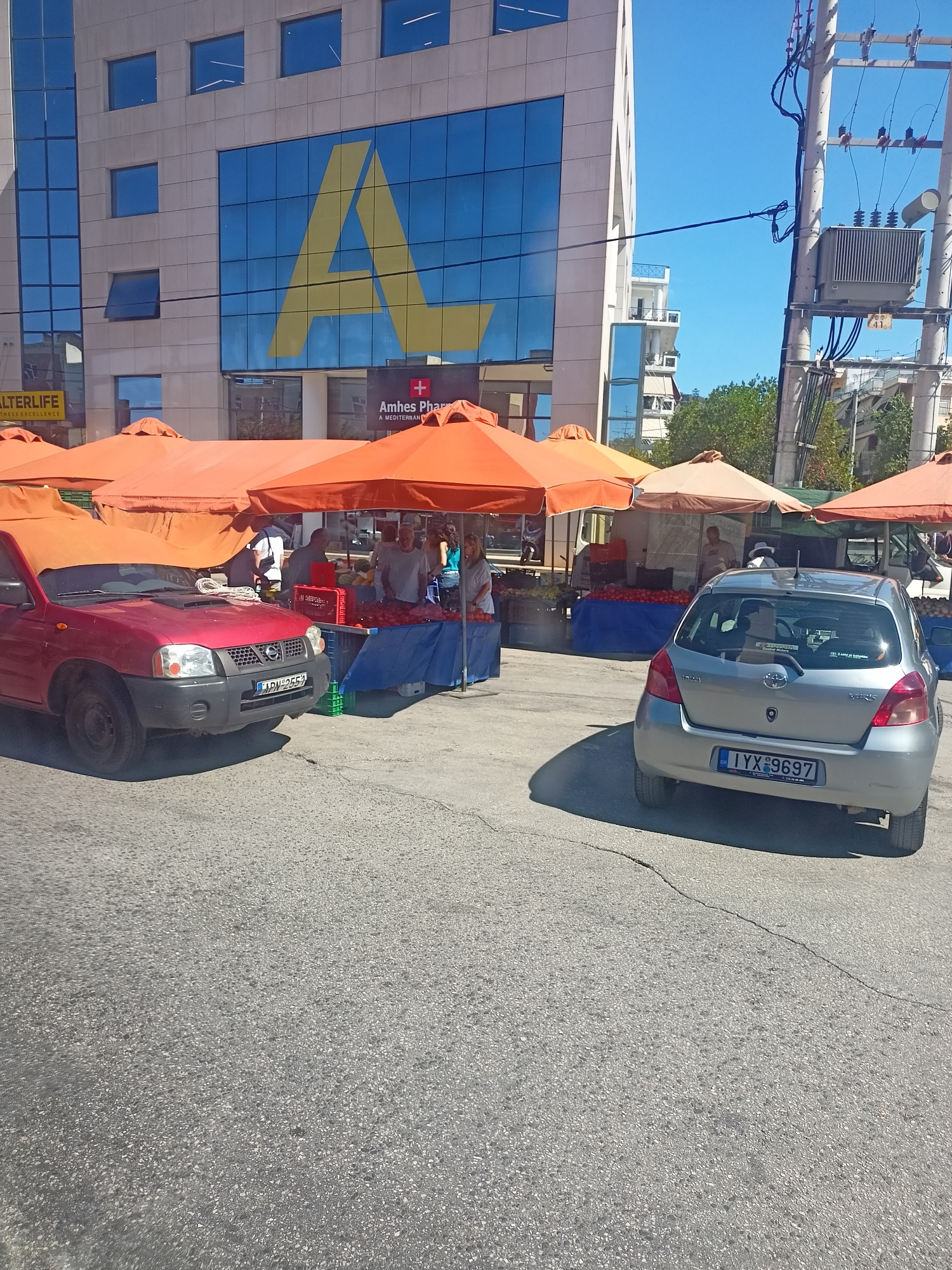 An outdoor market scene is captured on a sunny day in a city, possibly in Greece given the license plates. In the foreground, a red Nissan pickup truck is parked on asphalt. Behind it, a row of orange market stalls are set up, providing shade. These stalls display various fruits and vegetables, with vendors attending to customers. Several people are visible, some behind the stalls and others browsing. To the right of the stalls, a silver Toyota Yaris is parked. In the background, a modern building with large blue windows, featuring a prominent yellow "A" logo, stands tall. Utility poles and electrical equipment are visible on the right side of the image. The ground is paved with asphalt and shows some cracks.