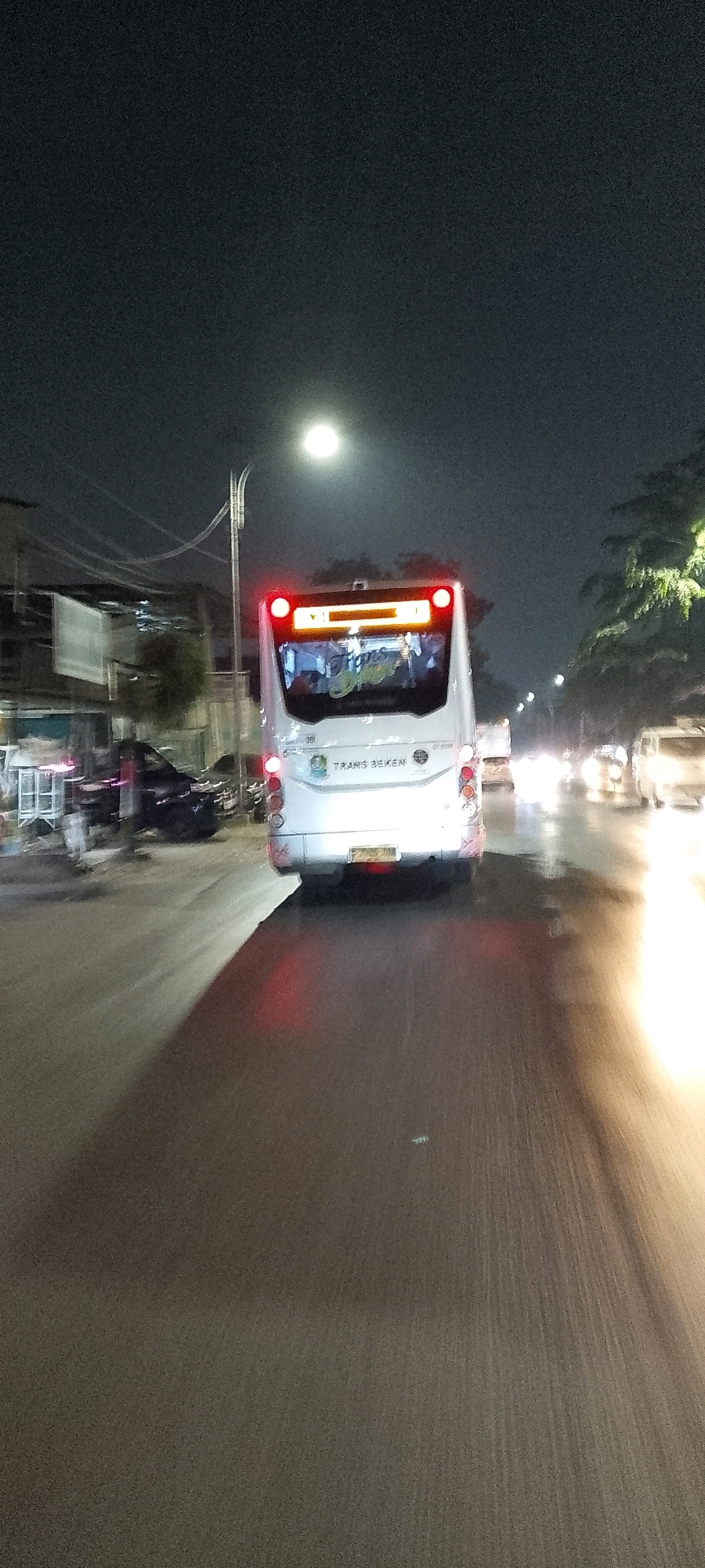 This is a low-light, motion-blurred vertical photograph taken at night in the city of Bekasi, Indonesia. The primary subject is the rear of a white bus labeled "TRANS BEKEN". The bus has its red taillights illuminated, and its license plate is visible, though illegible due to the blur. Above the license plate, there is an electronic display showing what appears to be route information or advertisements. The bus is in motion on a wet road, suggested by reflections of lights and the sheen on the pavement.
To the left of the bus, several other vehicles are visible, also blurred by motion, including what appears to be a black car and possibly a motorcycle. In the background, there are buildings with lights on and trees. A street lamp with a bright, circular light source is visible in the upper left quadrant of the image, contributing to the overall illumination of the scene. The sky is dark, indicating nighttime. The environment suggests an urban setting, likely a street or highway. The motion blur indicates the photograph was taken from a moving vehicle, likely another car or bus.