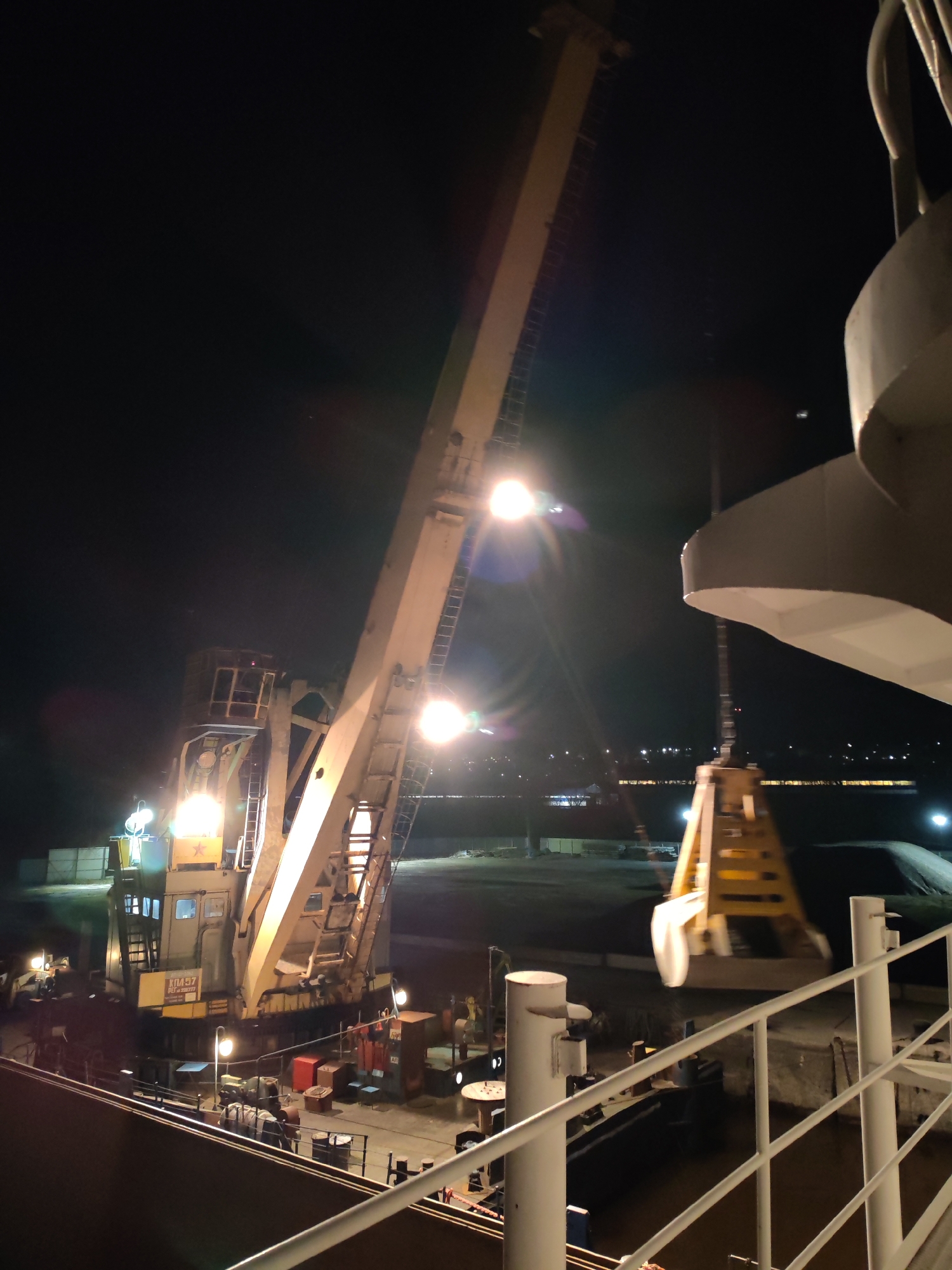 The image shows a nighttime scene, possibly at a port or industrial area. A large, illuminated crane is prominently featured, with its boom extended upwards. A grab bucket hangs from the crane, likely being used for loading or unloading materials. In the background, there's a glimpse of buildings or structures with lights visible. The foreground includes parts of a ship or platform, with railings and other equipment present. The location is likely in the city of Yakhroma, Russia, given the prompt. The overall setting suggests industrial activity, possibly related to shipping or construction.