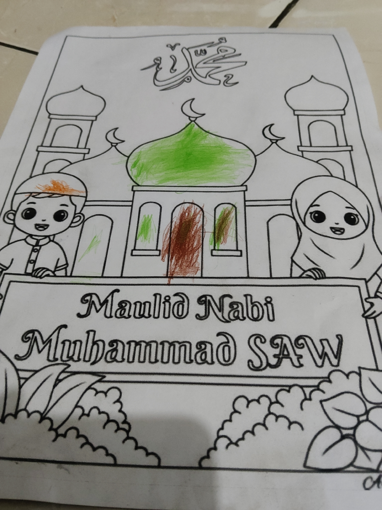 The image displays a coloring page featuring a mosque, two children, and Arabic calligraphy. One child, a boy, is on the left wearing a peci and a sarong-like shirt. The other child, a girl, is on the right wearing a hijab and a similar shirt. Both children have wide smiles. The mosque has domes and minarets. The calligraphy at the top reads "Muhammad". Below the mosque, the words "Maulid Nabi Muhammad SAW" are written, indicating the celebration of Prophet Muhammad's birthday. The coloring page shows some coloring applied: green on the central dome, orange on the boy's peci, and brown and green scribbles on the mosque's windows. Decorative elements like clouds and leaves are present at the bottom. The image is taken from a slightly elevated angle, with the coloring page lying on a patterned floor. The context of Mekarmukti, Indonesia, is not visually discernible from the image itself.