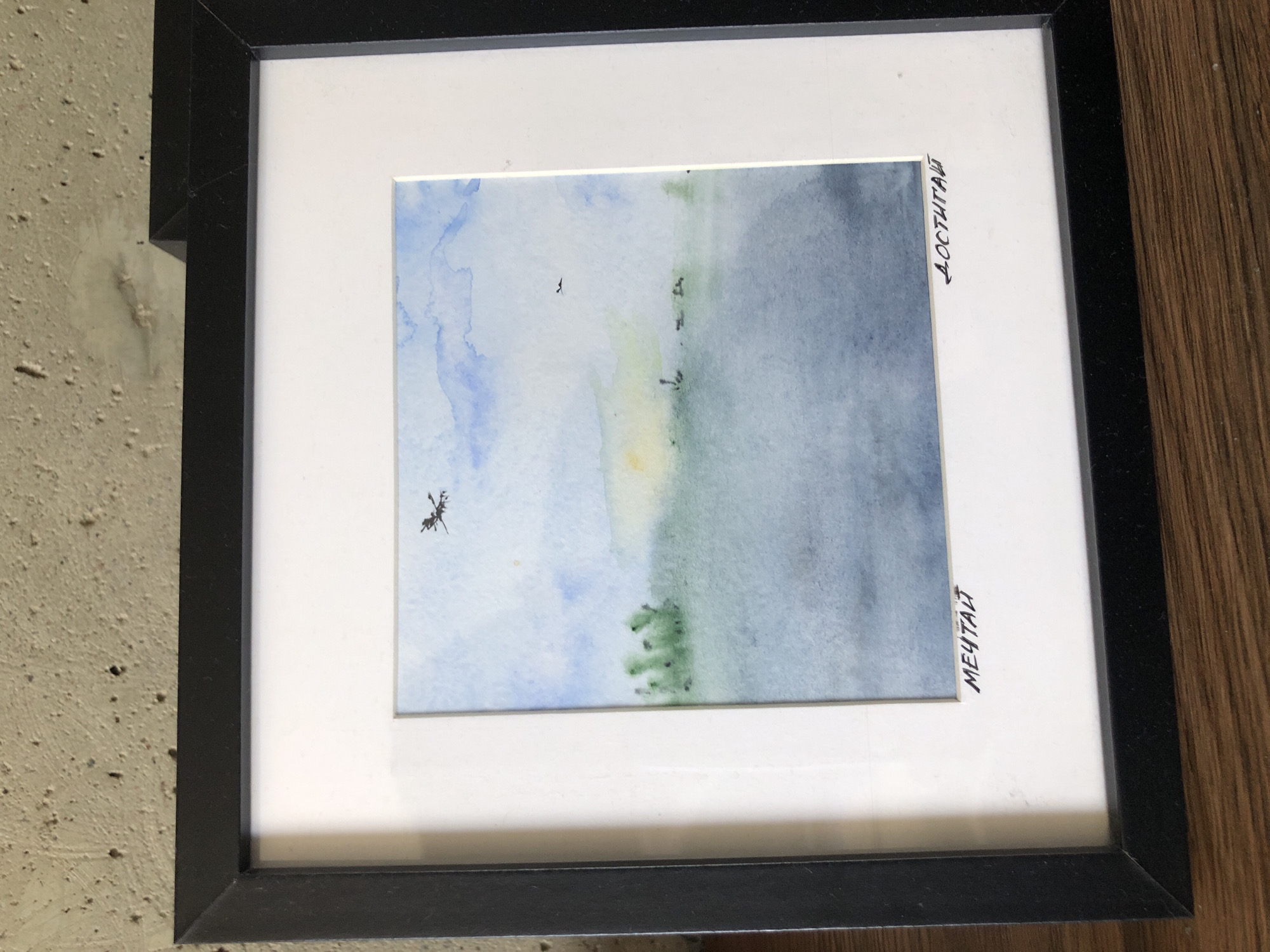 The image shows a framed watercolor painting depicting a serene landscape. The painting is matted with white cardstock and framed in black. The artwork features a body of water in the foreground, transitioning to a hazy green shoreline with sparse vegetation under a soft blue and yellow sky. Two small dark figures, possibly birds or insects, are visible in the sky. There are Cyrillic inscriptions in black ink along the bottom edge of the matting, on the left reading "МЕЧТАЙ" (Dream) and on the right reading "ДОСТИГАЙ" (Achieve). The painting is placed on a wooden surface, and the background suggests a concrete wall. No specific location in Saint Petersburg is identifiable from the visual information presented.