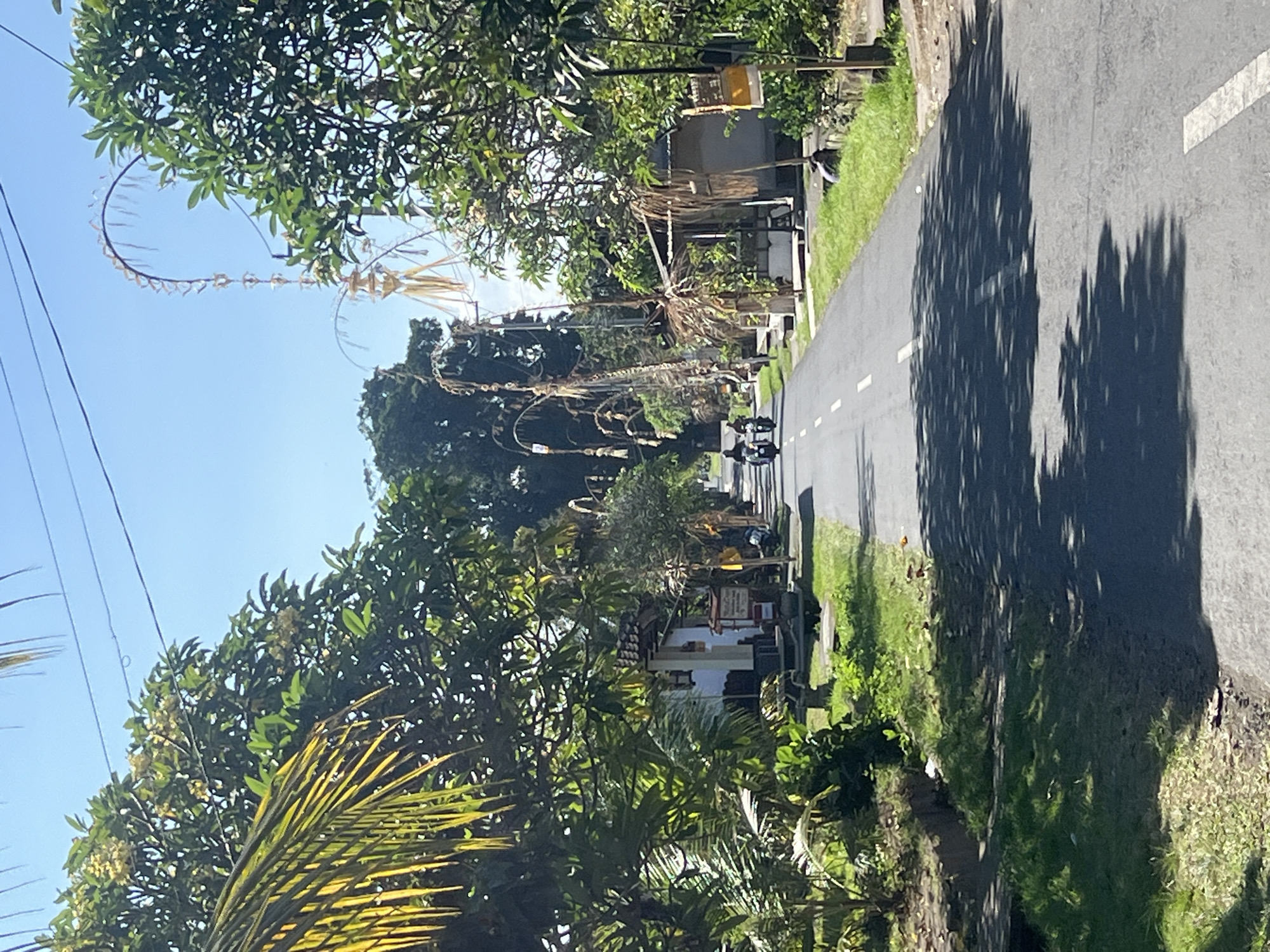 The image depicts a straight, asphalt road in the city of Sukawati, Indonesia, under a clear blue sky. Lush green vegetation and trees line both sides of the road, with some foliage casting shadows onto the asphalt. Several buildings, appearing to be small houses or shops with white walls and tiled roofs, are visible on the right side of the road. Traditional Balinese decorative elements, possibly Penjor, are prominently displayed on poles along the roadside, characterized by their tall, arched structures adorned with woven materials and palm leaves. A few individuals are seen riding motorcycles on the road, heading away from the viewer. The scene appears to be a typical street in a tropical, rural or semi-rural setting within Sukawati.