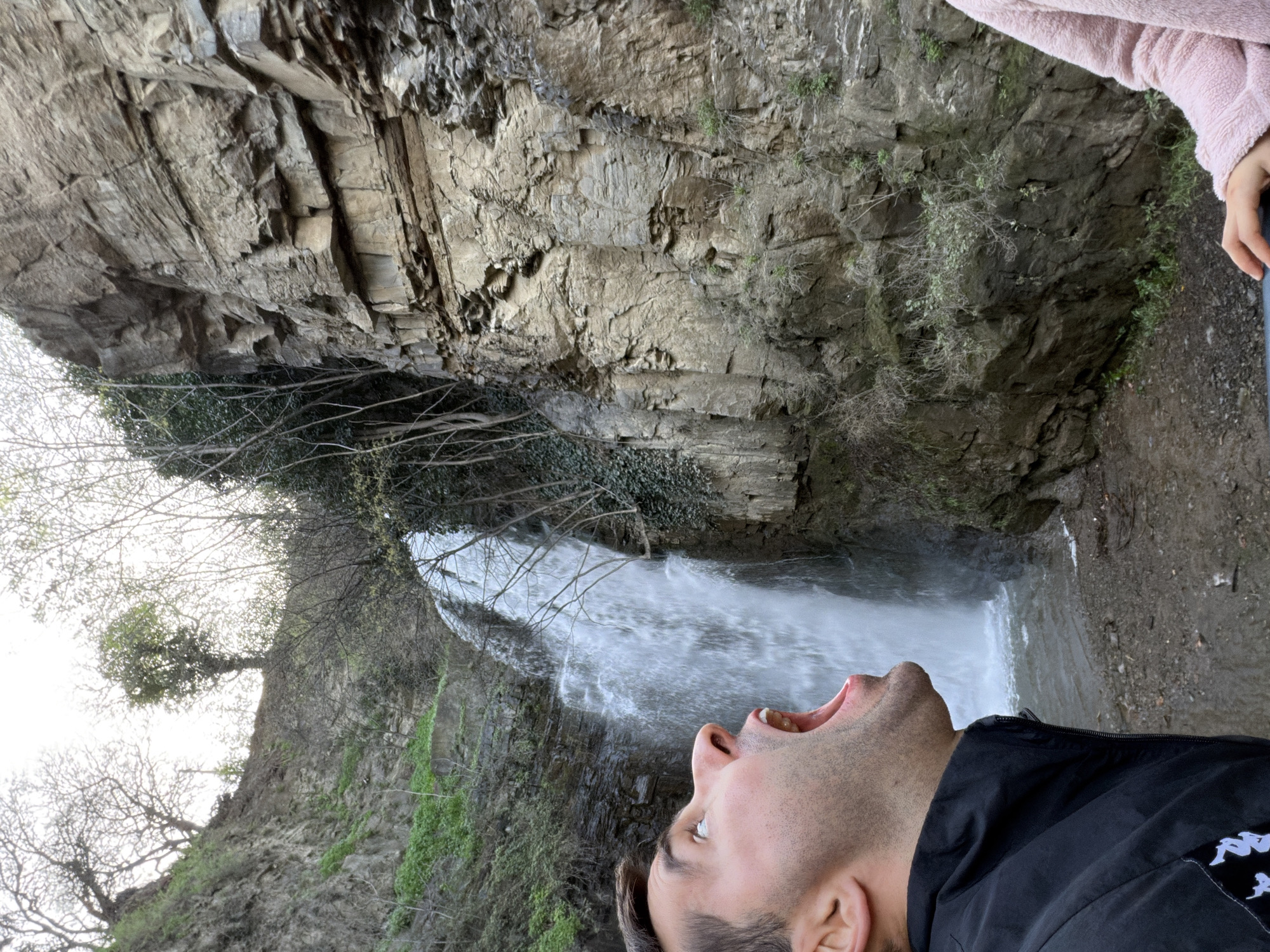 A man with dark hair stands near the base of a waterfall. He wears a black jacket, with his head tilted back and mouth open, seemingly trying to catch the water. The waterfall cascades down a rocky cliff face covered in sparse vegetation. Another individual wearing a pink sweater stands nearby. The location is Leghvtakhevi Waterfall, in the Old Town of Tbilisi, Georgia.