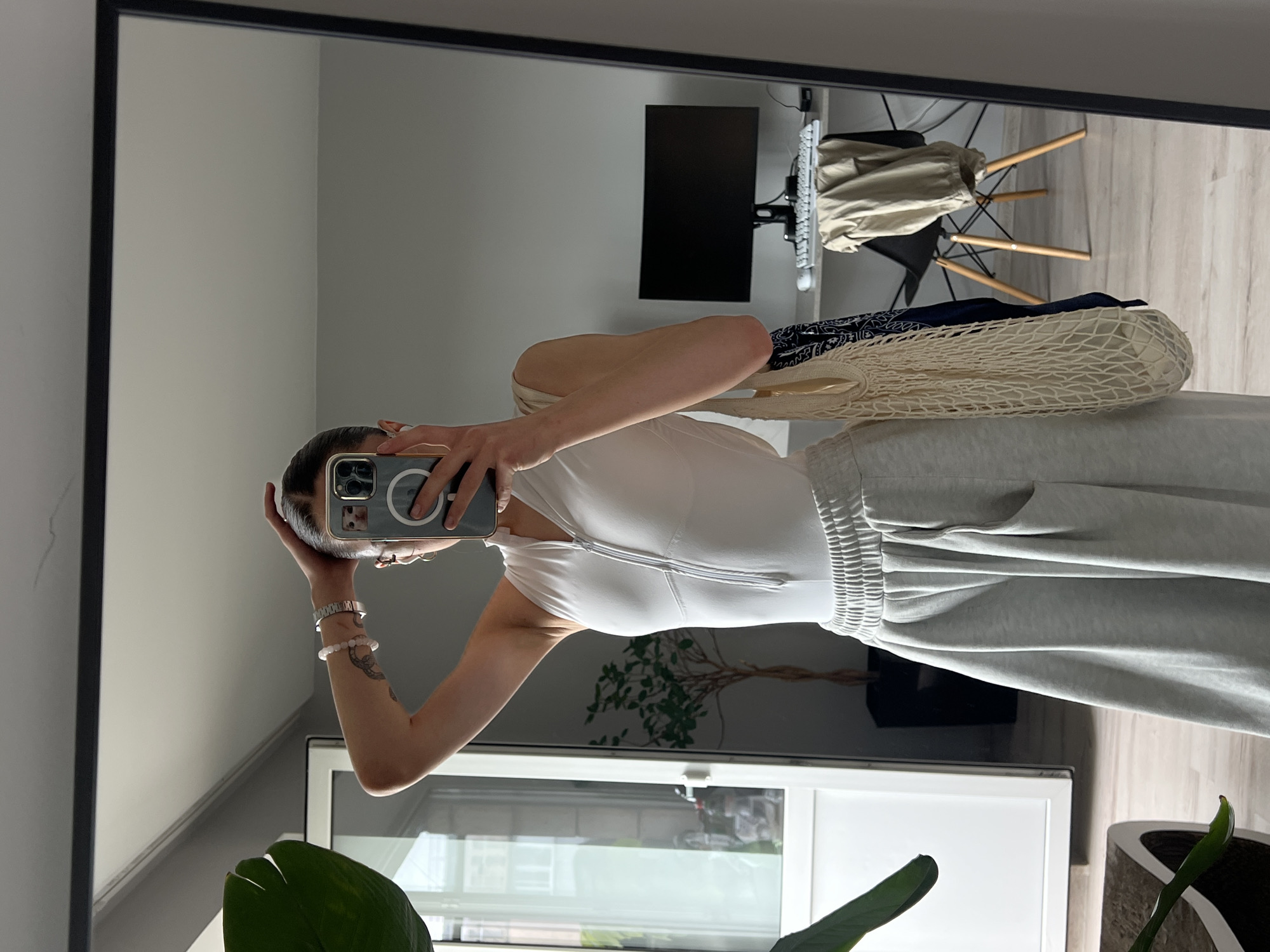 A person is taking a mirror selfie in an apartment, possibly in Ternopil, Ukraine. The person is wearing a white sleeveless zip-up top and light grey sweatpants. They are holding their phone to take the picture and have one hand on their head. A white mesh bag with a dark blue scarf inside hangs from their shoulder. In the reflection of the mirror, an office desk with a computer monitor, keyboard, and mouse is visible, along with a black chair with a beige jacket draped over it. A tall plant with green leaves is in the foreground on the left side of the image. A white door is visible in the background on the left.