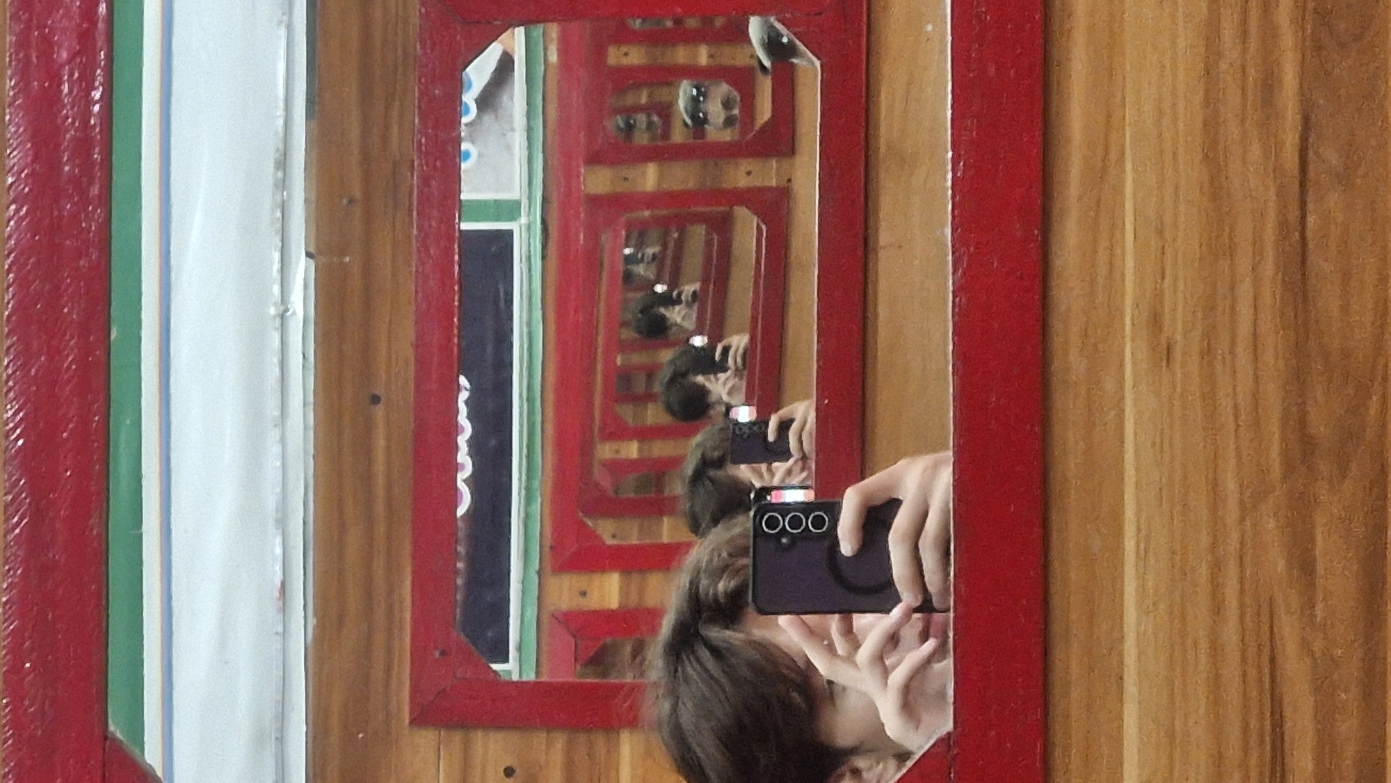 A person is taking a selfie in a mirror, holding up a black smartphone in their left hand. The smartphone has three visible camera lenses. The person's right hand is also visible, supporting the smartphone. The mirror is framed by a red border and is placed against a wooden surface. In the reflection, multiple instances of the person and the mirror arrangement are visible, suggesting a room with several mirrors or a reflection within a reflection. The background of the mirror shows a white surface with some colorful patterns and what appears to be text. The scene is indoors, with warm lighting and a focus on the reflective surfaces. The location is identified as Vashisht, India, suggesting it might be in a guesthouse, a shop, or a personal space within the town.