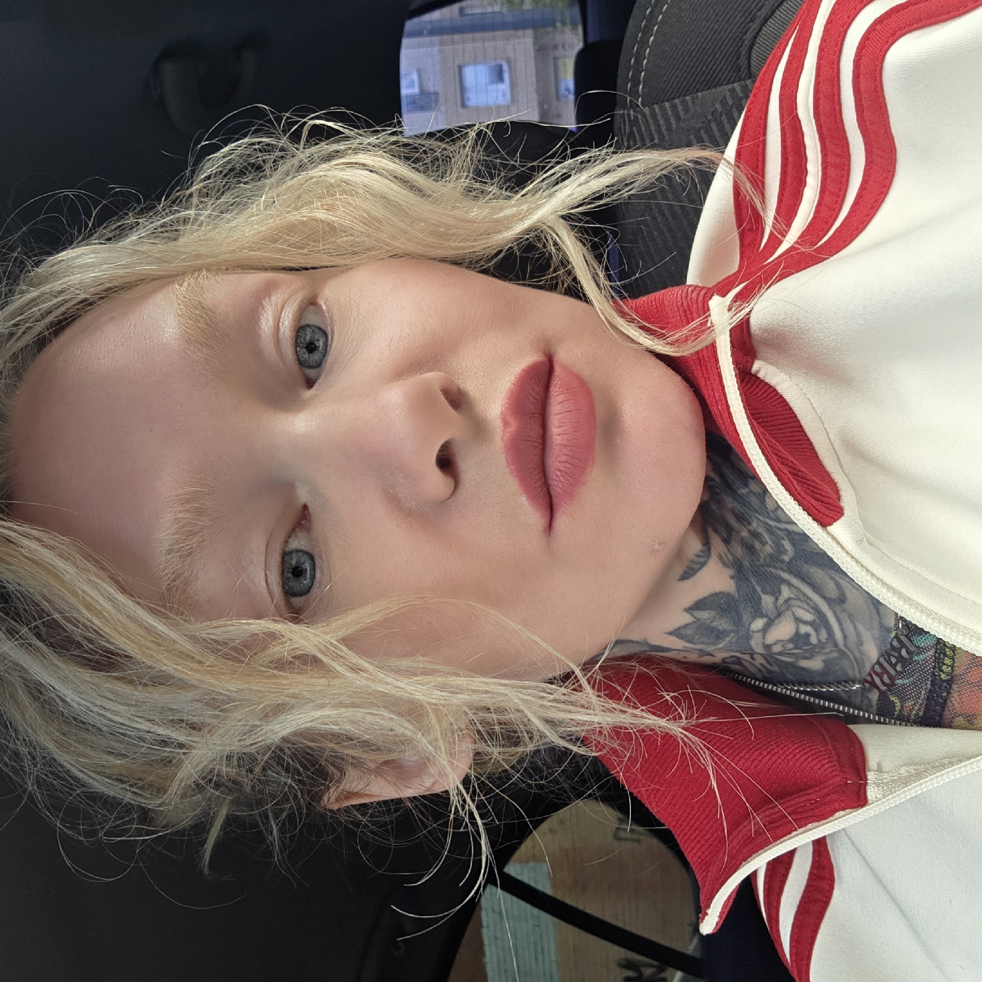 A fair-skinned individual with blonde hair, light eyes, and full, pink-toned lips is visible from the chest up. They are wearing a white jacket with red stripes and a red collar. A tattoo is visible on their neck. The individual is seated in a car. Outside, in the background, is a multi-story building. Based on the architectural style, the location could be in Saint Petersburg, Russia.