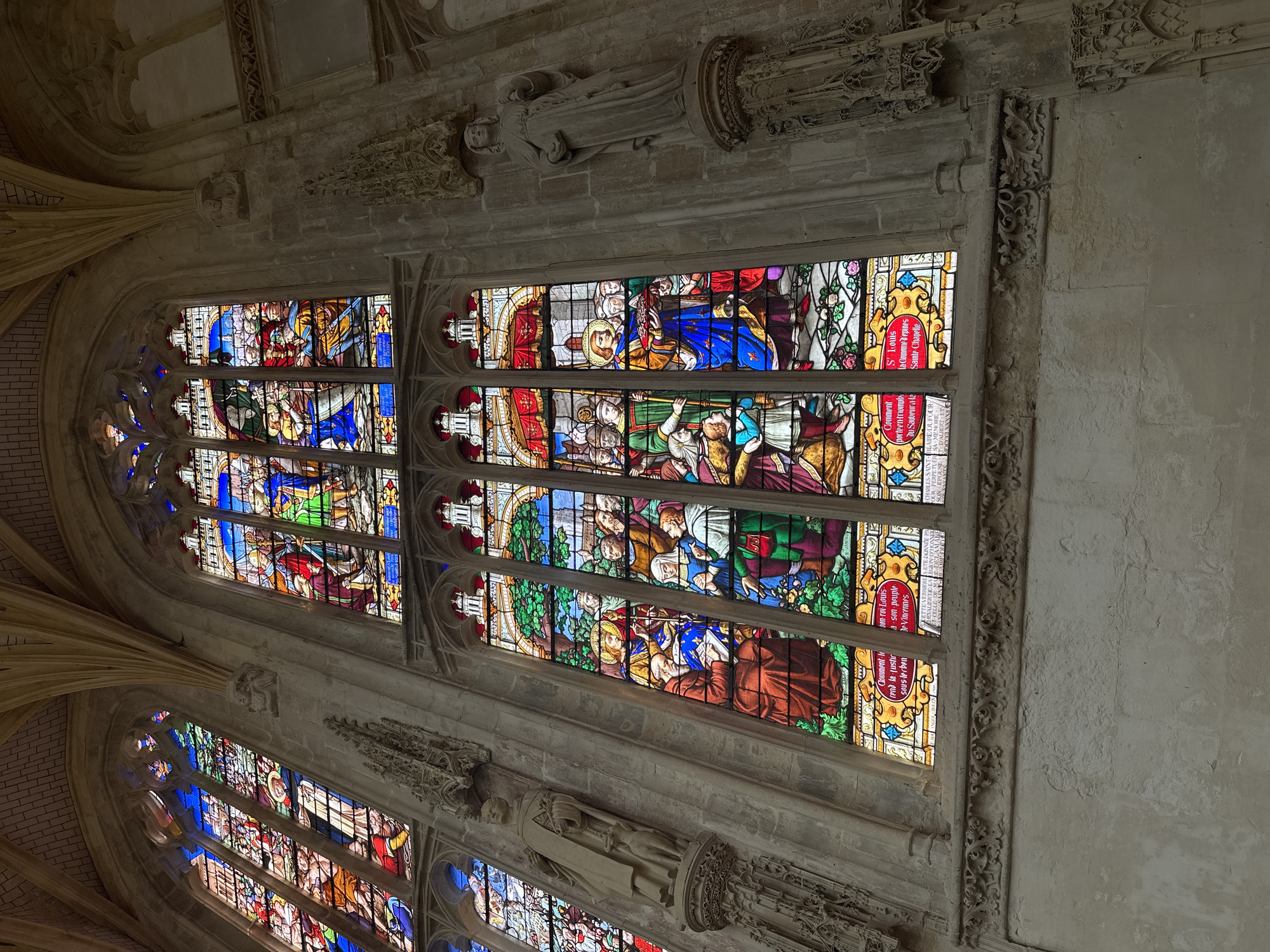 The image depicts the interior of a gothic church, likely Saint-Jacques Church in Dieppe, France. The focal point is a large, ornate stained-glass window. The window is divided into multiple panels, each depicting religious scenes rendered in vibrant blues, reds, greens, and yellows. Figures in historical or biblical attire are present, engaged in various actions such as gesturing, holding objects, and interacting with each other. Below the main window, a decorative stone frieze runs across the wall, featuring intricate carvings. Two stone statues of robed figures are positioned to the left and right of the window, attached to the stone columns of the church. The stonework of the church interior is rough and aged, with vaulted ceilings visible in the upper portion of the image. The lighting suggests an indoor, likely dimly lit environment, with light filtering through the stained glass.