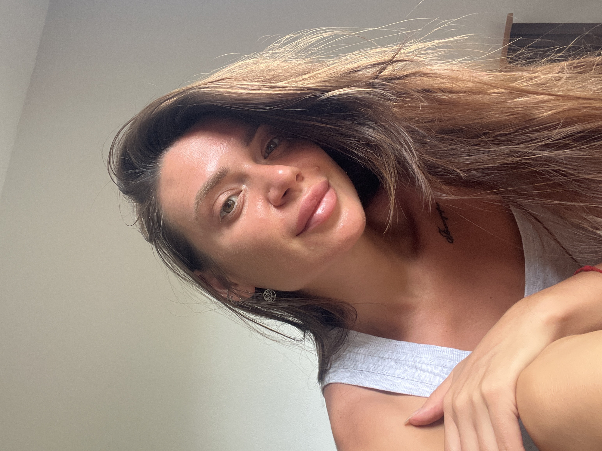 A woman with long, wavy, brown and blonde hair is pictured in a close-up, angled selfie. She has fair skin, prominent cheekbones, full lips, and light brown eyes. She is wearing a light gray tank top and a small silver earring is visible in her left ear. A tattoo is partially visible on her chest/collarbone area. Her right hand is gently placed on her right shoulder. The background is a plain, light-colored wall. The city and specific location are unknown.
