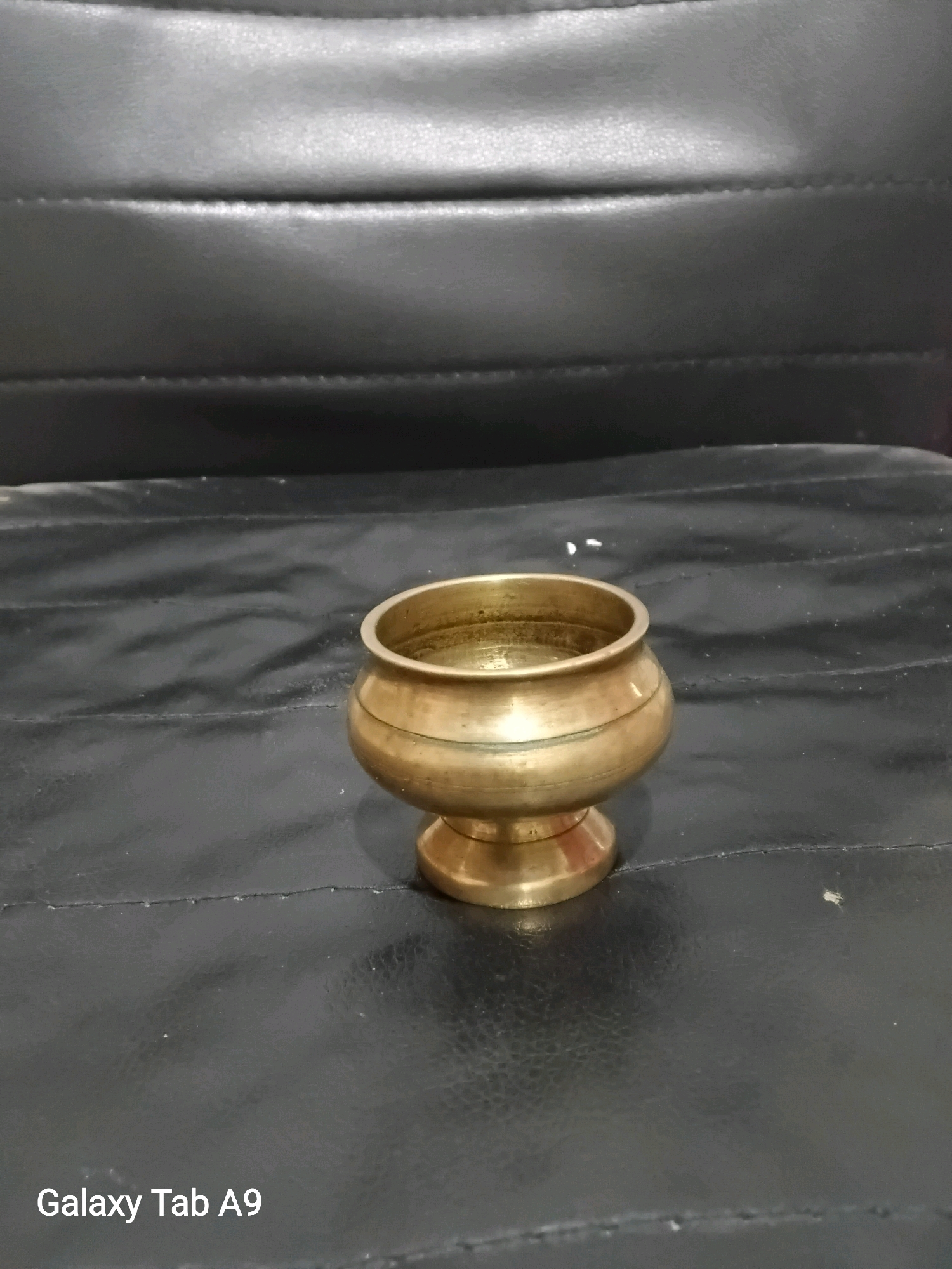 A small, antique brass bowl with a pedestal base sits on a black, textured surface. The bowl has a rounded body with two distinct horizontal bands. The surface appears to be a dark, possibly leather or vinyl material, with visible stitching lines creating a ribbed pattern. The background consists of a similar black, textured surface, which appears to be upholstery, possibly from a chair or sofa, with horizontal stitching. The lighting is overhead, casting subtle shadows. The lower-left corner of the image displays the text "Galaxy Tab A9". The scene is indoors and devoid of any specific urban or natural environmental cues to pinpoint a location in Karangan, Indonesia.
