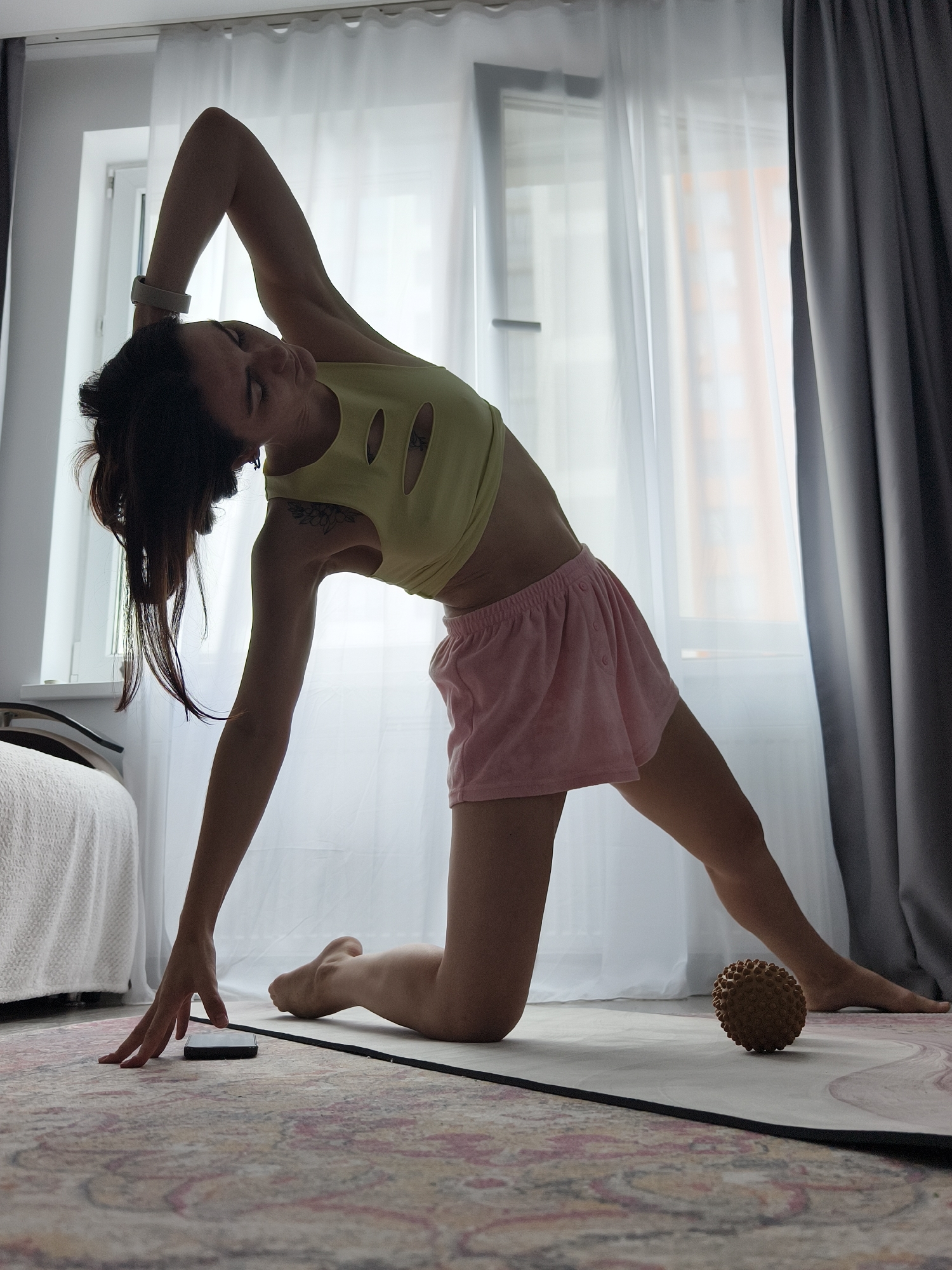 An adult female is depicted in an indoor residential setting, possibly within an apartment in the city of Tyumen, Russia, performing a side-bending stretch on a yoga mat. She is kneeling with her right leg extended to the side, her right foot flat on the mat, and her right hand touching a dark smartphone resting on a patterned rug. Her left arm is arched overhead, with a light-colored fitness tracker visible on her wrist. She wears a light yellow crop top with circular cutouts on the front and pink short shorts. A tattoo is visible on her upper left arm, and her dark hair is partially visible, appearing to be in an updo with some strands falling forward.
The room is illuminated by natural light entering from a large window, which is covered by sheer white curtains and a partially drawn dark grey curtain on the right. Through the sheer curtains, faint outlines of external urban buildings, including one with an orange hue, are discernible. To the left, a portion of a bed covered with a white textured blanket is visible. A spiky, light brown massage ball rests on the light-colored yoga mat, which features a subtle pink and white swirled pattern and is situated on a multi-colored patterned rug with hues of cream, pink, and grey.