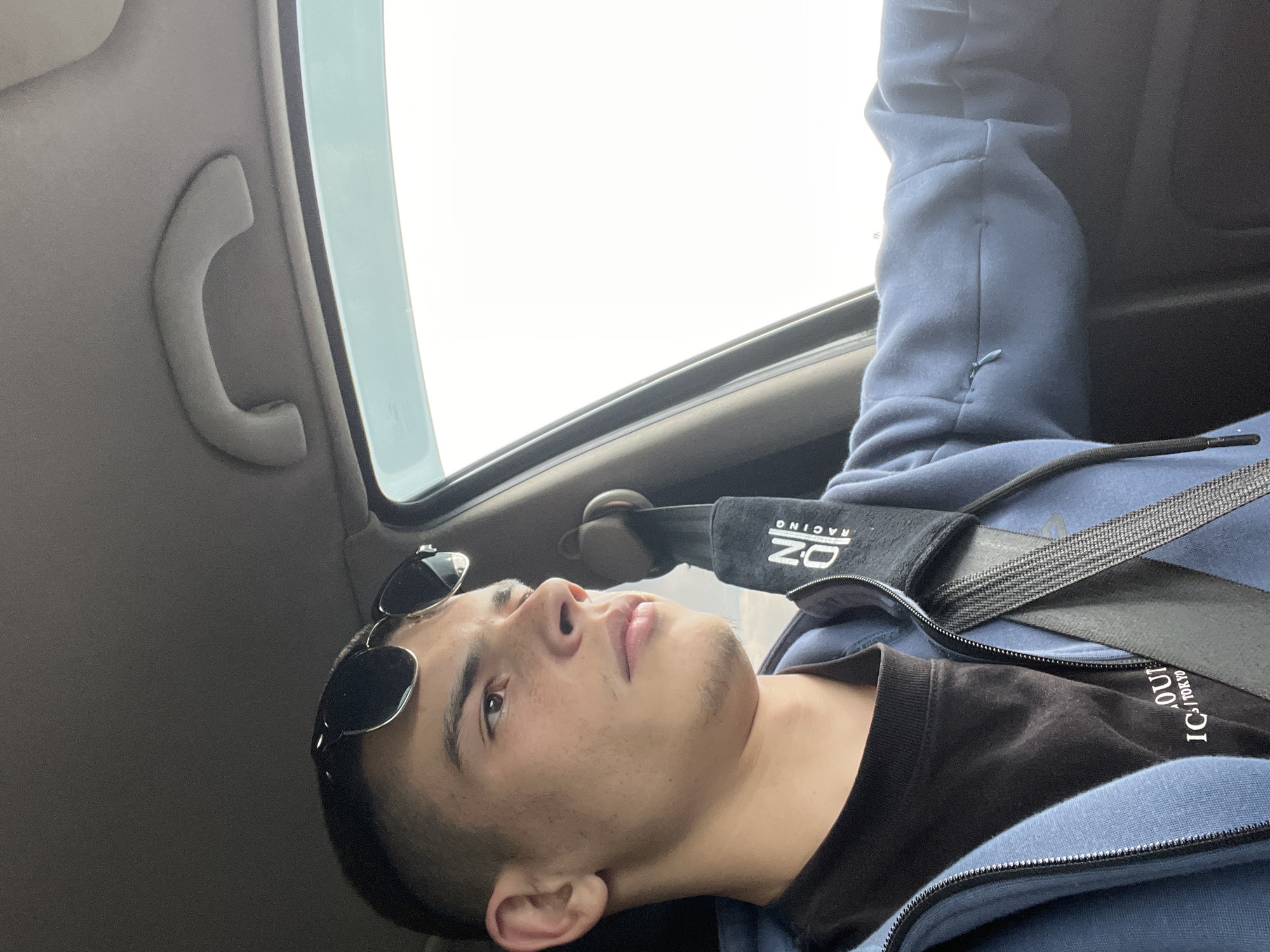 A young man is lying down in what appears to be the passenger seat of a car. He is wearing a blue hoodie and a black t-shirt, with sunglasses perched on his forehead. A seatbelt is across his chest, with an "OZ Racing" branded pad. The car's interior includes a grab handle visible near the window. The background outside the car is bright, suggesting daylight. The image does not contain enough information to establish a specific location in Dolna Mahala, Bulgaria.