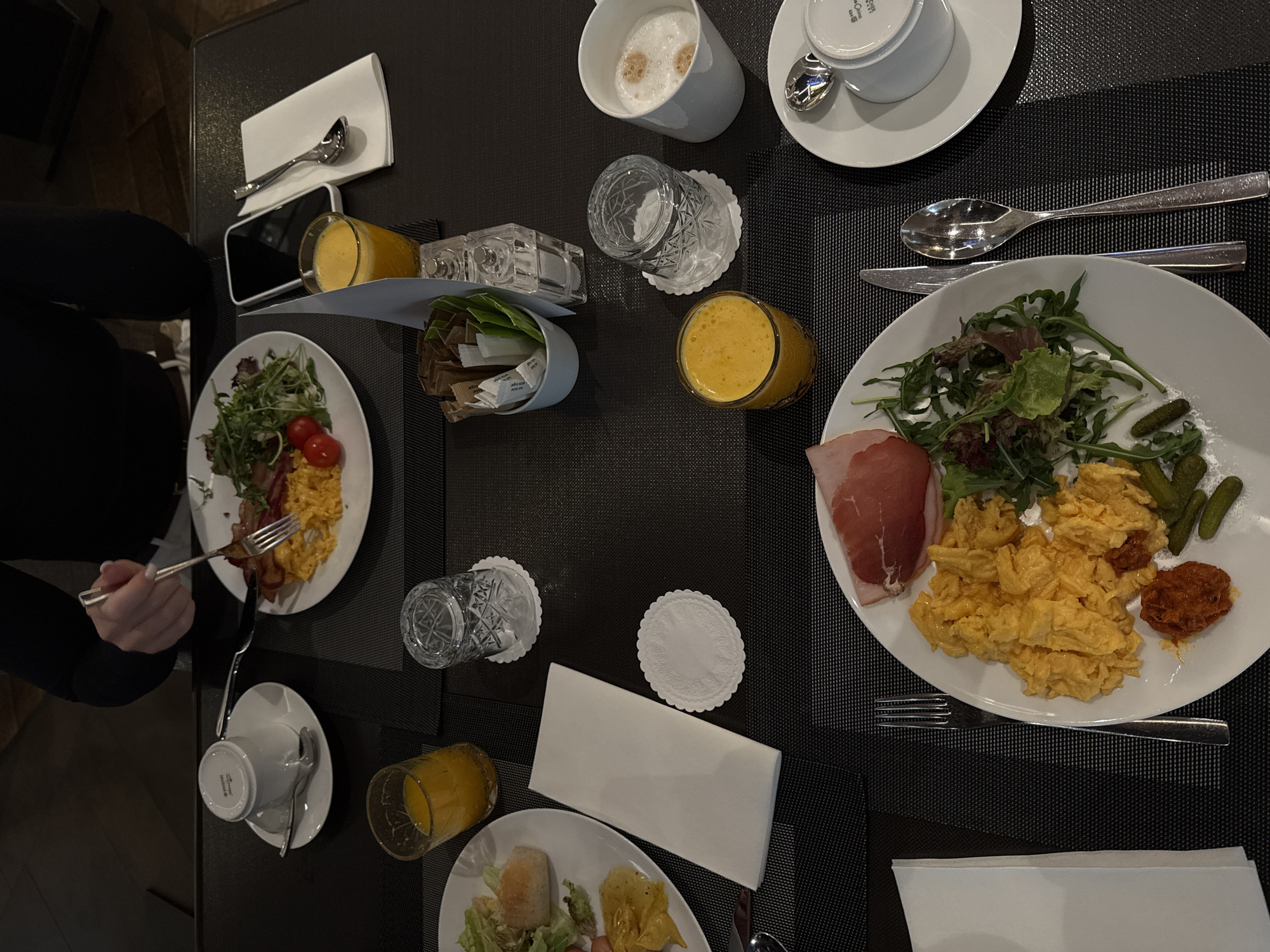 The image shows a breakfast table setting, likely in a hotel or restaurant in Vracar Urban Municipality, Serbia, given the presence of European currency, with multiple plates of food and drinks. A person in a black top is holding a fork and eating from a plate of scrambled eggs, bacon, and salad. There is another plate of similar food to the right. Other dishes on the table include orange juice, coffee, a container with sugar packets, a glass of water, napkins, and cutlery, all arranged on a dark-colored table. A phone also sits near a plate of food.